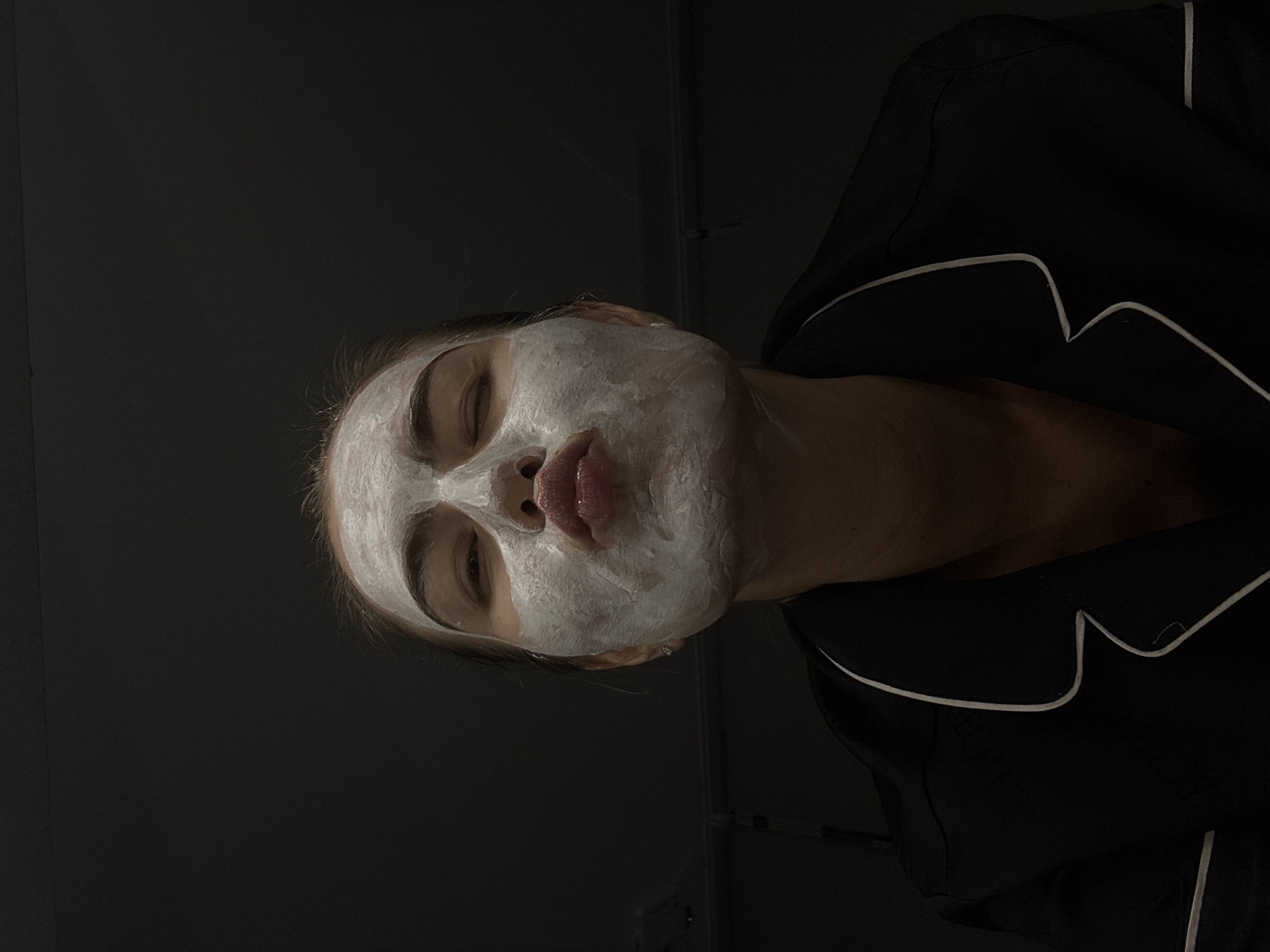 A woman in Unknown, Unknown is depicted from a low angle, giving the viewer a perspective from below her face. She has a white facial mask applied, covering her entire face, except for her eyes, and mouth, which is pursed slightly. She is wearing dark pajamas with a light-colored trim around the collar. The background is dark, adding to the focus on her face.