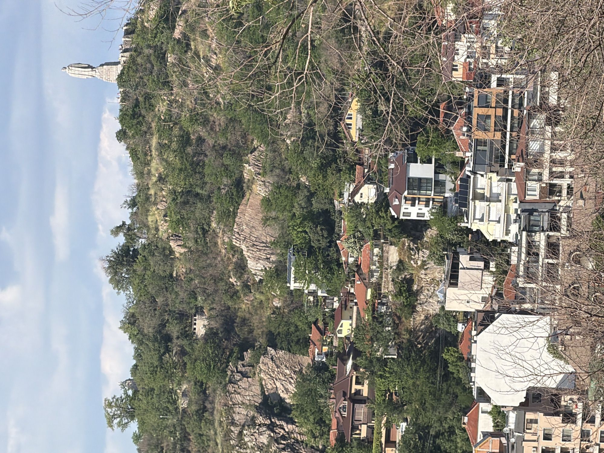 An aerial shot shows the city of Plovdiv, Bulgaria, with the Alyosha Monument atop Bunardzhik Hill prominently displayed on the left. The shot reveals a mix of residential architecture, nestled amidst lush greenery and rocky terrain. The Alyosha Monument, a large stone figure, stands against a backdrop of sky and scattered clouds. The architecture in the scene ranges from traditional red-roofed buildings to modern, multi-story constructions. A network of tree branches partially obscures the view in the foreground. The overall scene suggests a blend of urban development and natural landscape, characteristic of Plovdiv.