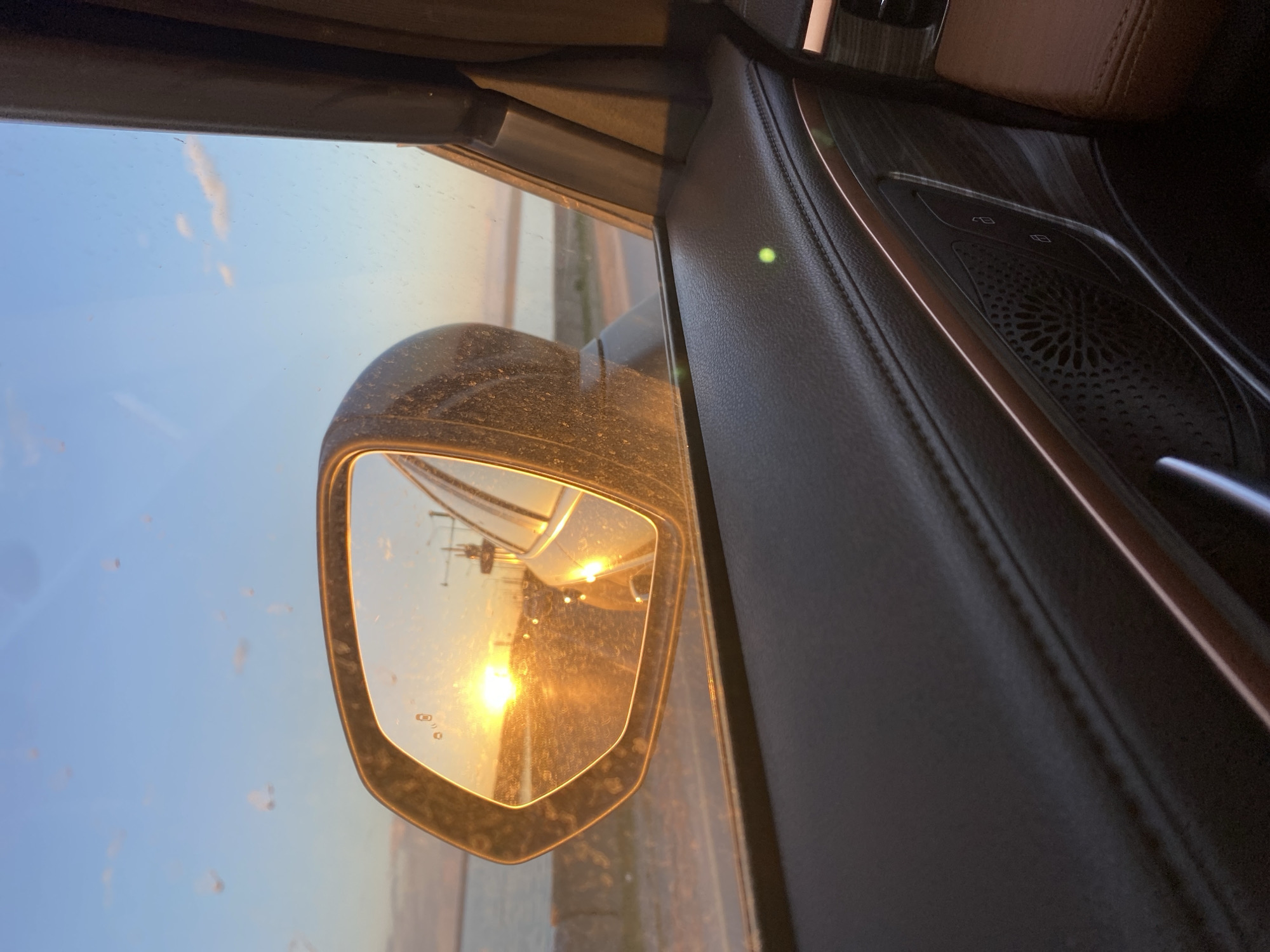 The image is an interior view from a car, looking out at the side mirror. The mirror reflects a sunset or sunrise over what appears to be an industrial area. The sky is blue fading to orange near the horizon. In the reflection, there is a structure that could be a crane or utility pole. The mirror itself appears to be dusty. The car's interior features black leather trim with brown accents on the panel and interior door, speaker grilles and power windows are visible. I lack the information to accurately determine whether the image was taken in Chaikovsky, Russia, or its location.