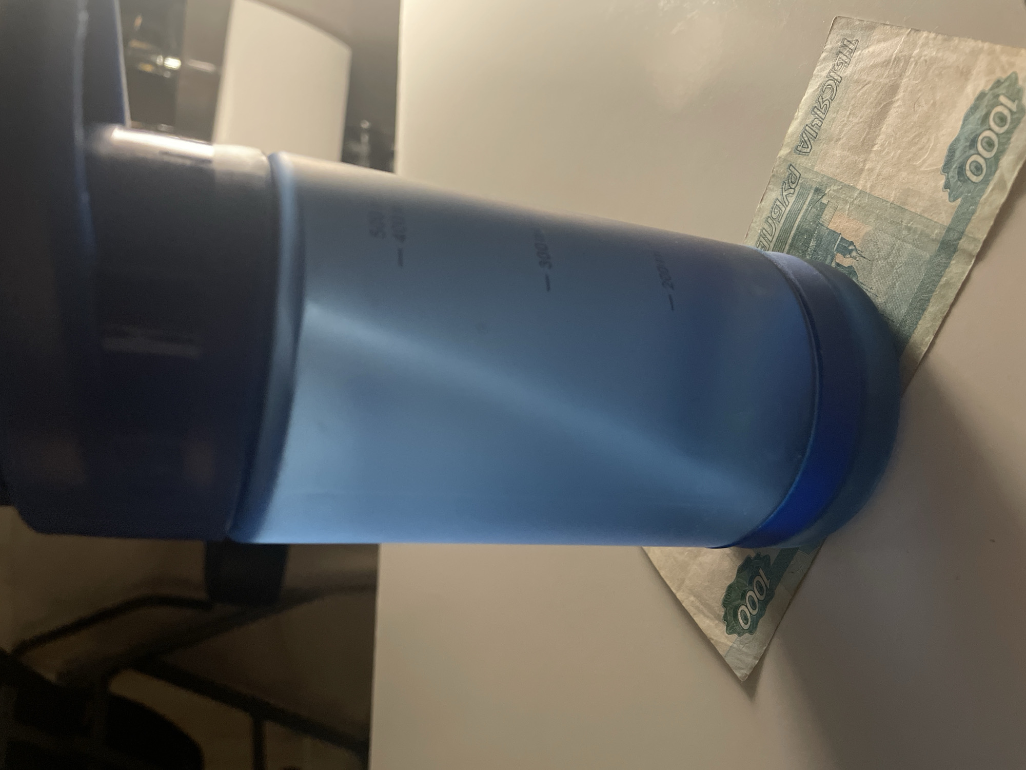 The image shows a close-up, horizontal shot of a blue translucent water bottle resting on a light-colored, flat surface. A folded Russian banknote is tucked under the bottle, with the denomination "1000" clearly visible in green numbers. The banknote also displays text in Cyrillic characters. The water bottle has measurement markings along its side, with "500ml", "400ml", "300ml", and "200ml" visible. The background is blurred, showing hints of a dark, possibly indoor environment with some light sources. The lighting in the image appears to be artificial and comes from the right, casting subtle shadows. The focus is on the bottle and the banknote, suggesting a potential demonstration of the bottle's capacity relative to the currency's value, or simply a snapshot of these items in proximity. There are no people visible in the frame. The overall impression is that of a still life arrangement.