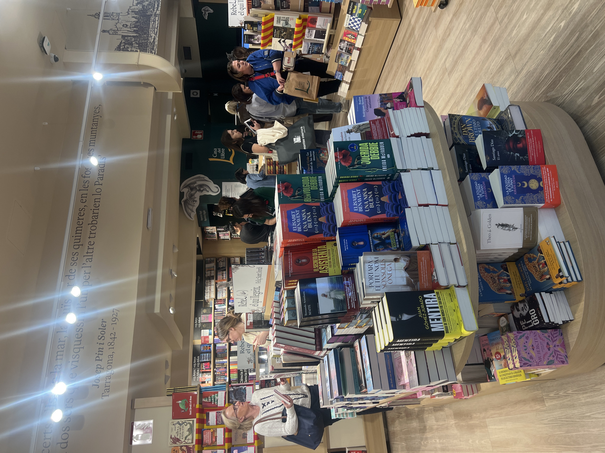 The image depicts the interior of a bookstore in Tarragona, Spain. A multi-tiered circular display unit showcases a variety of books, with titles in both Spanish and Catalan. Customers browse the selection, with some individuals examining books and others walking past. A text excerpt from Josep Pin i Soler, Tarragona, 1842-1927 is visible on the wall. The bookstore is well-lit with ceiling fixtures.