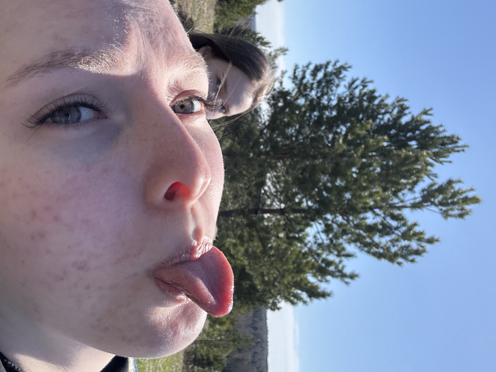A close-up portrait shows a fair-skinned person, likely female, sticking out their tongue towards the camera. She has light eyes, freckles, and visible eyelashes. Slightly behind her and to the right, a second person is partially visible with a similar fair complexion and darker hair. The background features green trees against a clear, light blue sky, suggestive of an outdoor setting. Based on the prompt's request for location, this photo is supposed to be taken in the city of Zhigalki, Russia, although this cannot be definitively confirmed from the image alone.
