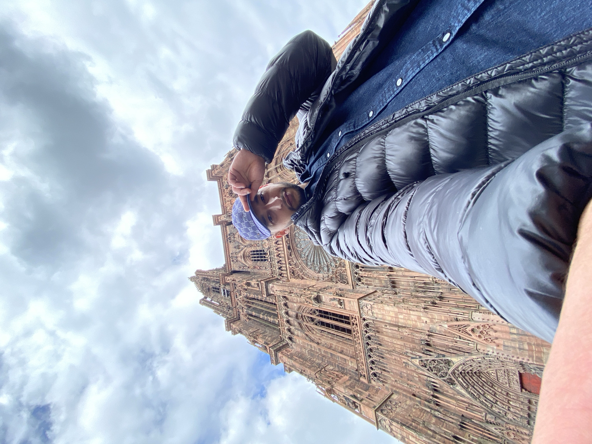 A man wearing a blue paisley bandana and a black puffer jacket with a denim shirt underneath stands in front of the Strasbourg Cathedral in Strasbourg, France. The cathedral is tall, ornate, and constructed of red sandstone. The sky is cloudy. The man appears to be taking a selfie.