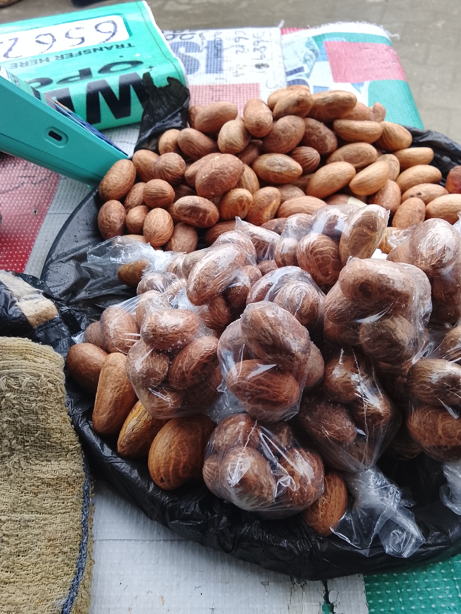 This is a close-up, overhead shot of a market stall in Lekki, Nigeria. The primary subjects are a large pile of nuts, some loose and others packaged in clear plastic bags. The nuts are oval-shaped with a rough, brown textured surface. They are arranged in a black plastic bag, which is part of the market stall setup. To the left, a portion of a burlap sack is visible, adding to the market ambiance. In the background, a turquoise-colored object with "TRANSFER HERE" and numbers "6567" printed on it is partially visible, possibly a point-of-sale device or a sign. Other patterned fabrics and a white surface are also present, suggesting the setting is an outdoor market. The lighting appears natural, indicating it's daytime. There are no people visible in the frame, focusing entirely on the goods for sale. The scene is typical of a Nigerian street market, showcasing local produce and retail practices.