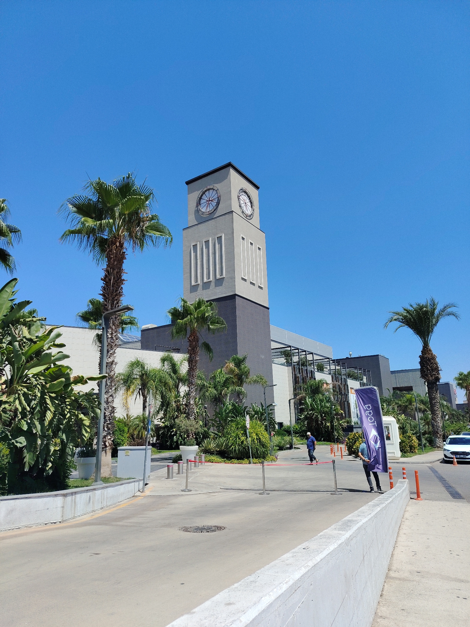 The image depicts the exterior of a modern building featuring a prominent clock tower, situated in Kepez, Turkey. Palm trees and lush greenery surround the structure, suggesting a warm climate. The building's architecture is contemporary, with a mix of light-colored panels and darker, possibly concrete, sections. The clock tower itself is tall and rectangular, with a clock face visible at its apex. In the foreground, a paved road leads towards the building, with bollards and temporary barriers indicating an entrance or checkpoint. A purple flag with white lettering stands near a small booth. Two individuals are visible near the entrance, one walking away from the camera and another facing it. A white car is parked on the right side of the road. The sky is clear and blue, indicating a sunny day.