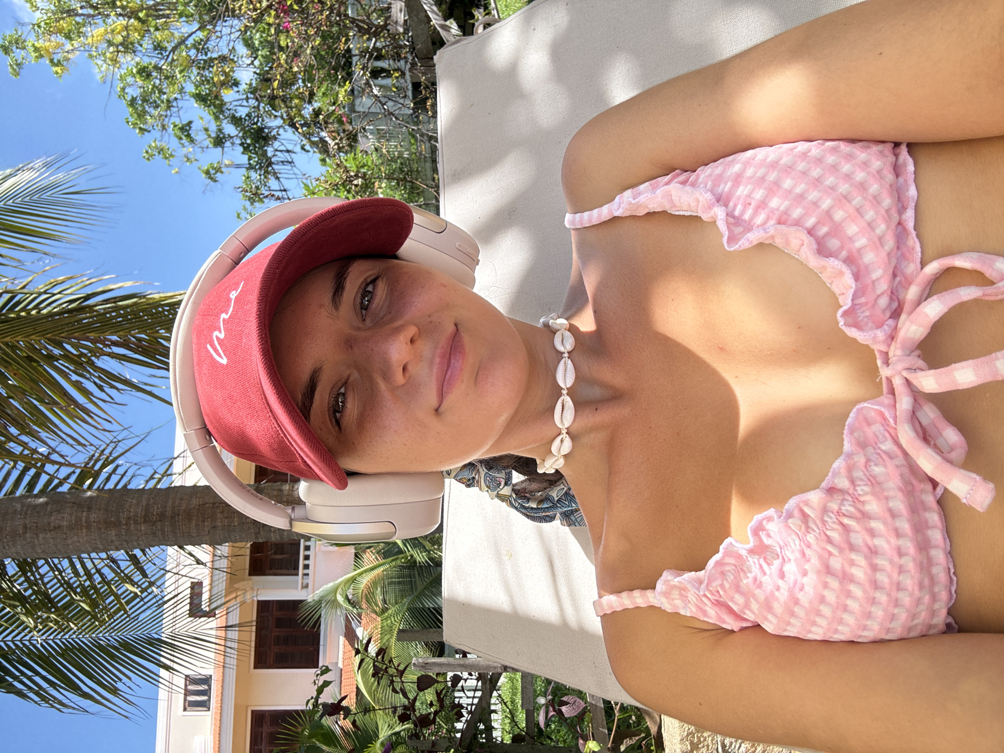 A woman wearing a red baseball cap, white headphones, and a pink gingham bikini top with a shell necklace is sitting on a lounge chair outdoors. She is looking at the camera with a slight smile. A palm tree and a building are visible in the background, suggesting a tropical or resort location. The scene appears to be set in Phu Thuy Ward, Vietnam.