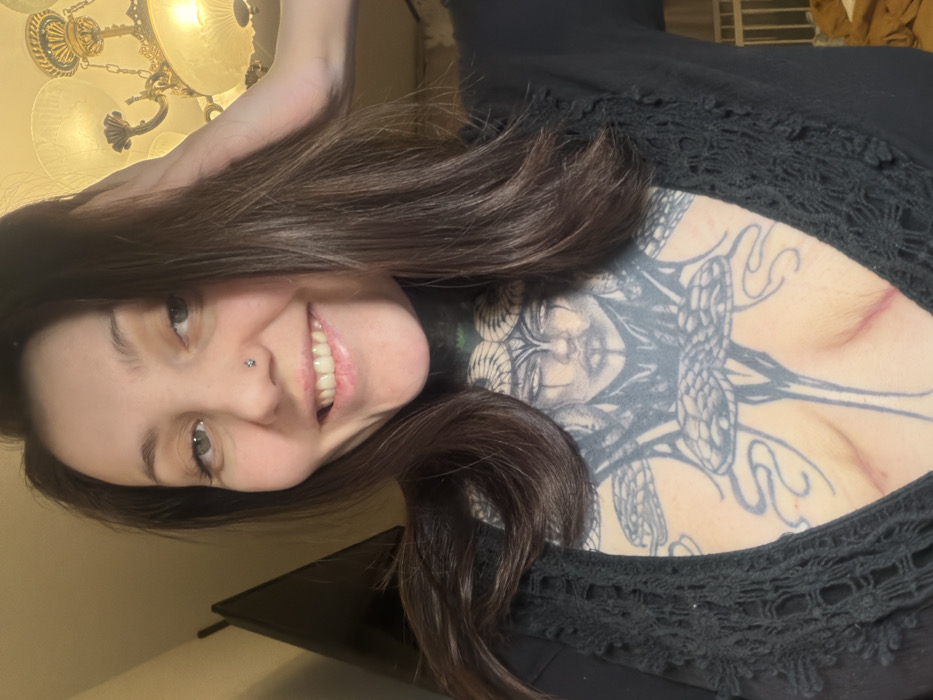 A woman smiles with a nose ring and long brown hair, likely taking a selfie. She wears a black lace-trimmed top or dress. The background includes a chandelier and a dark surface, possibly a table. Notably, she has a large tattoo on her upper chest or back depicting Medusa. The image is rotated.