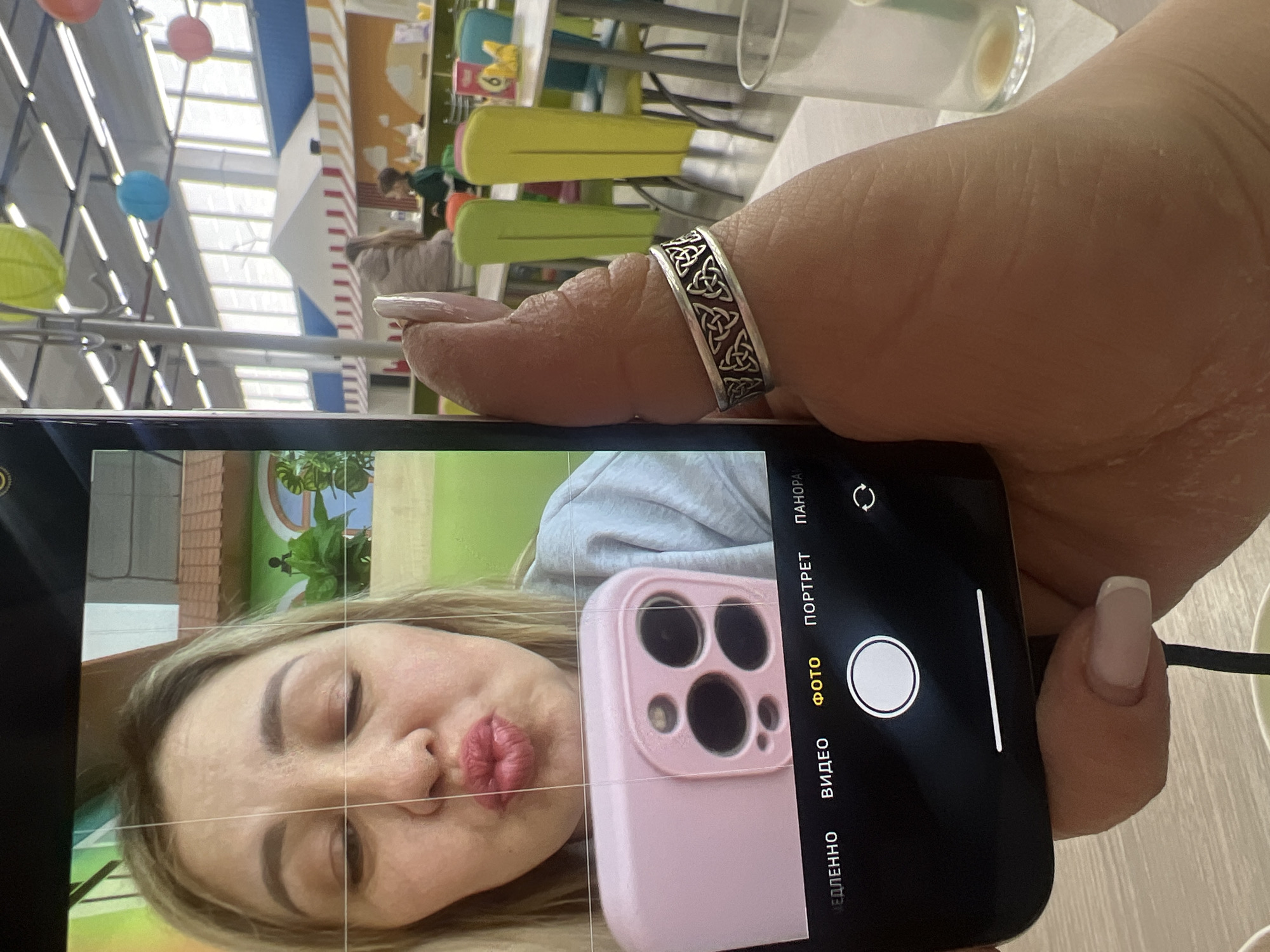 A person holds a black smartphone vertically in their right hand, capturing a selfie using the front-facing camera. The phone's screen displays a live camera preview of a woman with blonde hair making a "kissy face" with pursed, dark pink lips and closed or semi-closed eyes. She is wearing a light grey top. Within the selfie preview, her left hand is visible, holding a pink-cased smartphone with a triple camera array. The phone held by the primary subject features a camera interface with "ФОТО" (Photo) mode selected.
The hand holding the phone has light-toned skin, manicured nails with a French tip style, and wears a silver-colored ring adorned with Celtic knot patterns on the ring finger.
The background, largely out of focus, reveals a brightly lit indoor environment consistent with a children's entertainment center or family cafe. This location is identified as the **Funny Park children's entertainment center, located at Primorsky Boulevard, 26, Chaykovsky, Perm Krai, Russia, 617762**. The space features a high ceiling with exposed structural elements and fluorescent lighting. Colorful paper lanterns (green, blue, orange/pink) hang from the ceiling. In the mid-ground, small tables with brightly colored chairs (yellow, green, blue) are visible. Structures resembling stylized houses or market stalls with striped awnings (red and white, orange and white) decorate the walls. Large windows are present high on the far wall, admitting natural light. Blurred figures of other individuals, including an adult in a light jacket and a child in a green shirt, are present in the background. A clear glass, partially used, rests on a table in the bottom right corner of the frame.
