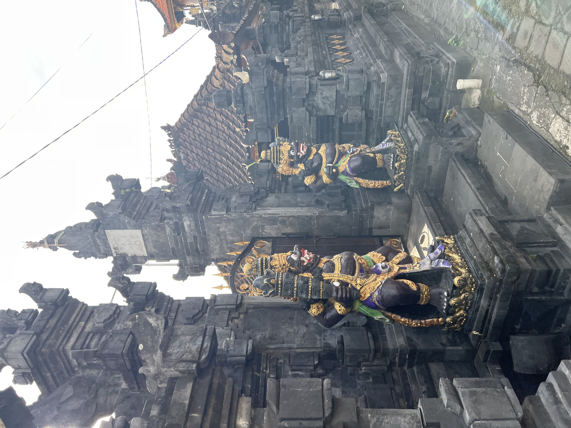 The image shows the entrance to a building in North Kuta, Indonesia, with traditional Balinese architecture. Two large, dark-colored statues of fierce guardian figures, adorned with gold and colorful accents, stand on either side of the entrance steps. The statues are intricately detailed, with open mouths and menacing expressions. The building itself is constructed of dark stone blocks, with ornate carvings and a tiled roof. The sky is bright and overcast. The scene appears to be a temple or a traditional Balinese dwelling.