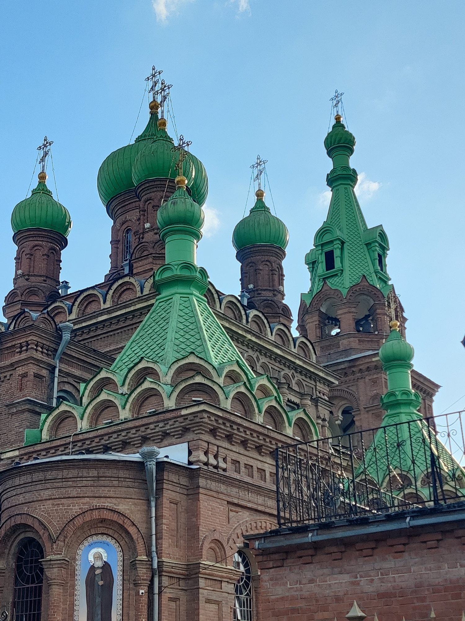 This is a medium-shot, eye-level, vertical, outdoor photograph taken on a sunny day with a clear blue sky, possibly in the morning or afternoon, as evidenced by the bright lighting. The image features a close-up view of the intricate architecture of a red brick church, characterized by multiple green domes and decorative elements.
The main subjects are the various domes of the church, each topped with a gilded Orthodox cross. The domes vary in size and shape, with some being rounded and bulbous, while others are more conical and steeply pitched. The red brickwork of the church walls is visible, showing arched windows and decorative cornices. One of the walls features a tiled mural depicting a robed figure with a halo, possibly a saint.
A black wrought-iron fence and a portion of a balcony are visible in the foreground, suggesting a viewpoint from a nearby elevated structure or walkway. The overall scene conveys a sense of architectural grandeur and religious significance. The location is identified as Krasnodar, Russia, which aligns with the architectural style of the church. No specific activity or event is depicted; the image focuses solely on the structure and its details. There is some text visible on the mural, but it is not legible in this resolution.