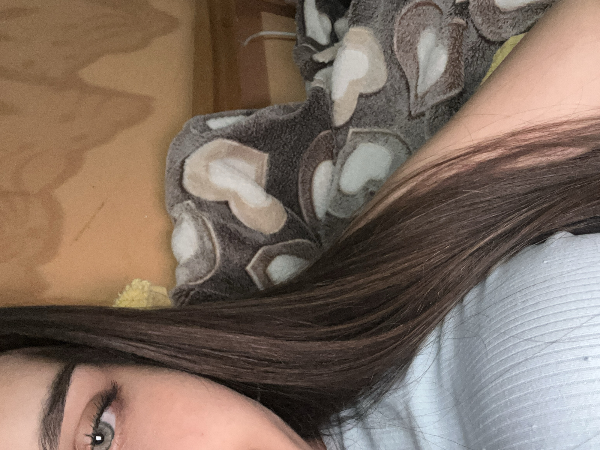 The image is a close-up, cropped view of a person's left eye, part of their face, hair, and arm. The person has light-colored eyes and dark brown hair. They are wearing a light blue ribbed top. In the background, a blanket with a heart pattern is visible, and behind that, a wooden surface. The lighting suggests an indoor environment, possibly a home. There is no discernible city or location information present in the image.