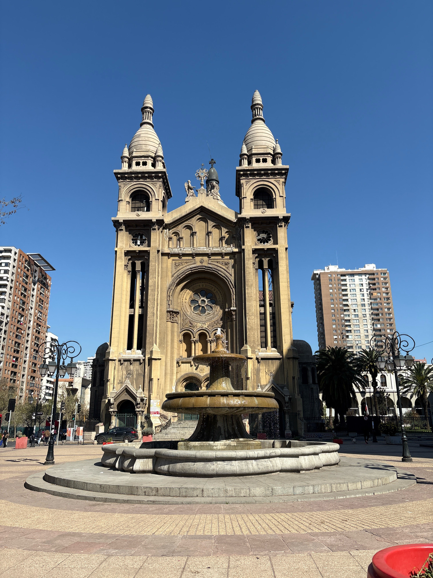 The image shows the Metropolitan Cathedral of Santiago, Chile, located in the Plaza de Armas. The cathedral is a grand, neoclassical building with two prominent bell towers topped with spires. In front of the cathedral, a multi-tiered fountain with water cascading down is visible. The plaza features a paved walkway with a textured pattern. To the left of the cathedral, a tall, modern residential building with numerous windows stands out against the clear blue sky. To the right, more trees and buildings, including another residential tower, are visible. Several people are walking in the plaza, and a black car is parked near the entrance of the cathedral. The scene is bathed in sunlight, suggesting a bright, clear day.