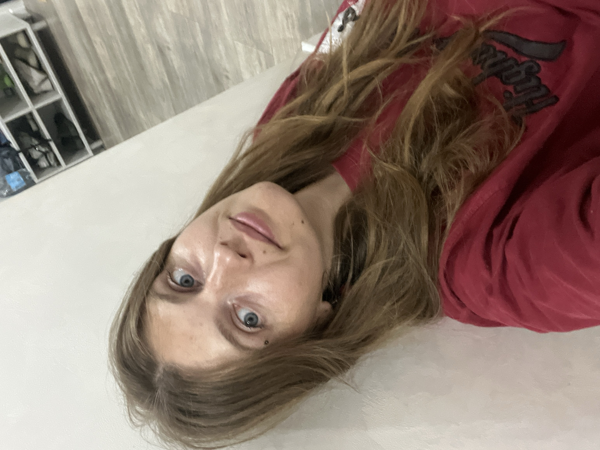 In an indoor setting, a young woman with long, wavy brown hair is leaning against a white wall. She is looking towards the camera with a slight tilt of her head and her body. Her eyes are blue, and she has a small piercing above her left eye. She is wearing a dark red long-sleeved shirt with a graphic on the front that appears to say "Highs". The background shows a floor with a wood-like pattern and a white shelving unit filled with shoes and other items, consistent with a storage area or entryway. The lighting is diffuse, suggesting an interior environment. The scene is captured from a slightly high angle.