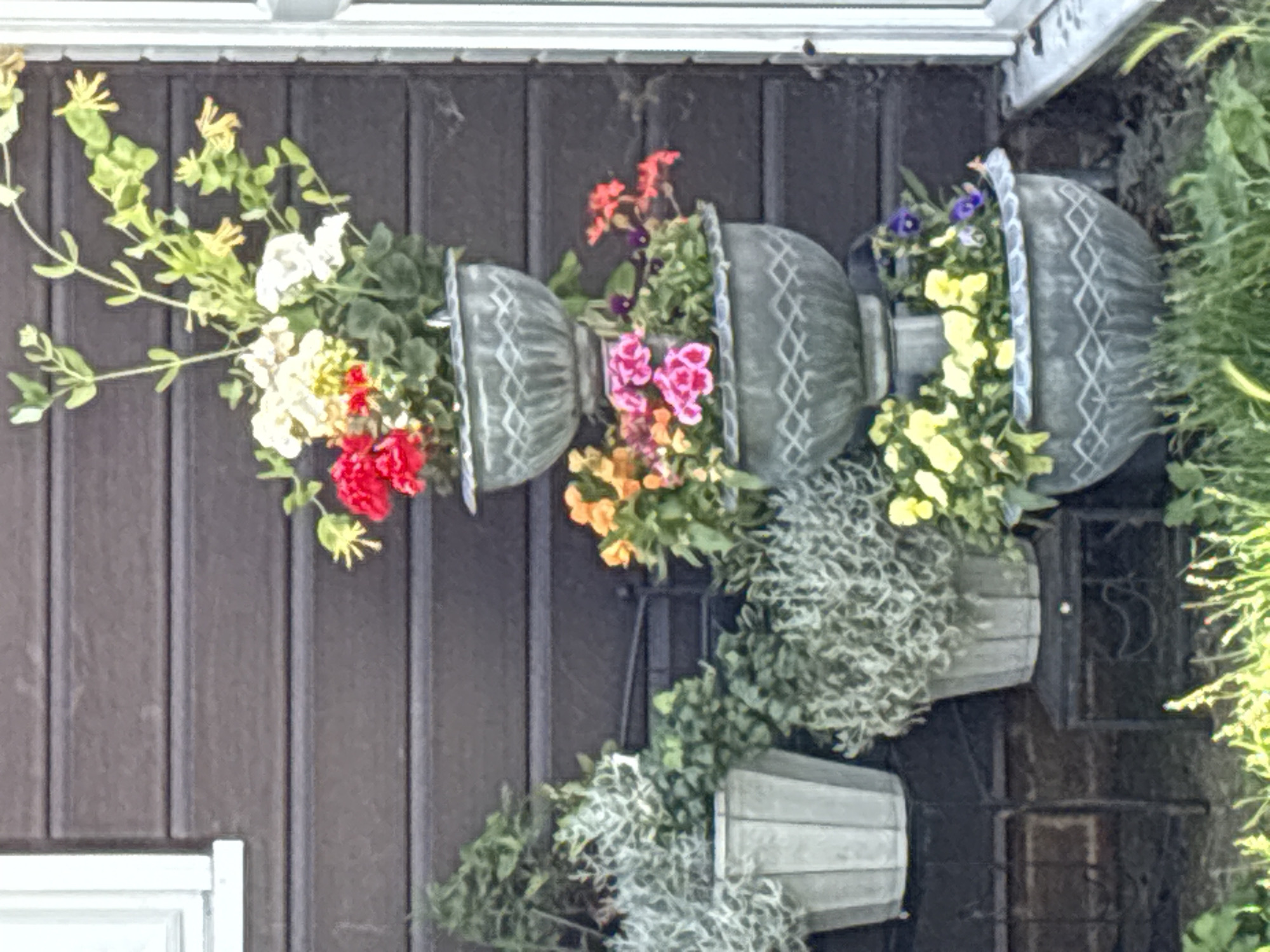 A tiered planter filled with flowers and foliage stands against a dark brown, horizontally planked wall. The planter is composed of three grey, ornate bowls stacked on top of each other. The top bowl contains red, white, and yellow flowers, along with green foliage. The middle bowl has pink and orange flowers and greenery. The bottom bowl is filled with yellow flowers, purple pansies, and green leaves. To the left of the tiered planter, another grey planter with silvery-green foliage sits on a black metal stand. The bottom portion of the image shows green grass and other plants. A white-framed window is visible in the upper left corner of the image.
