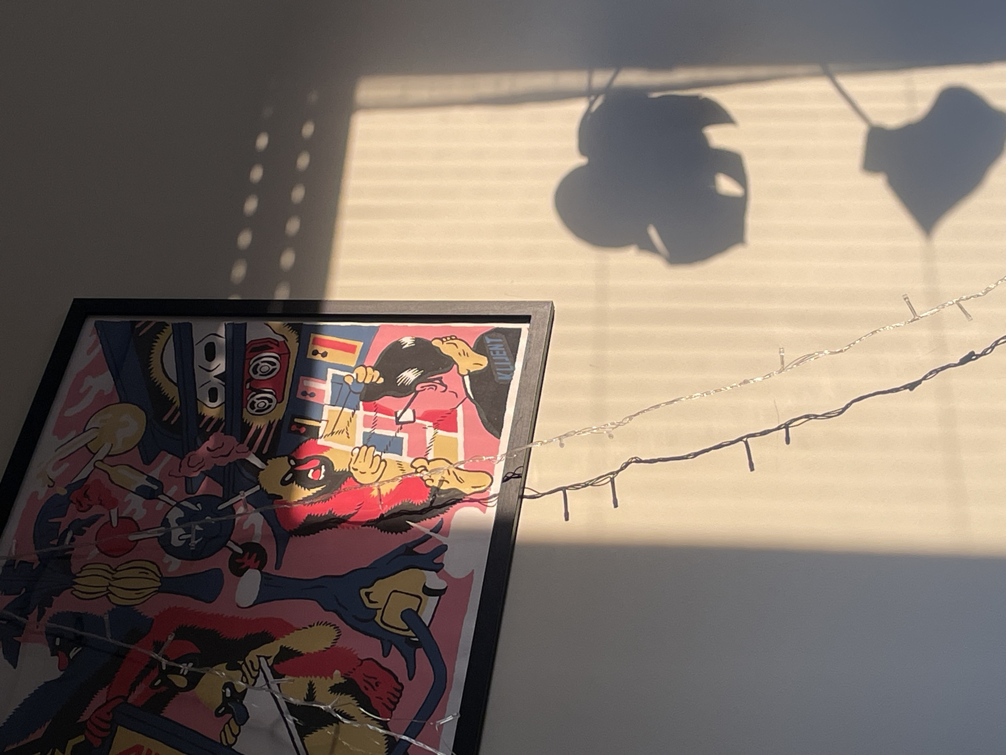 The image shows a framed art print leaning against a wall. The print features a vibrant and chaotic design with multiple figures and abstract shapes in colors like pink, red, yellow, and black. The print's lower portion is partially obscured by a string of small decorative lights.
The wall behind the print is off-white and illuminated by sunlight casting shadows of what appears to be plant leaves and window blinds. The light creates a stark contrast between the sunlit areas and the shadowed portions of the wall. The scene seems to be indoors.
There is no indication of the image location being in Vracar Urban Municipality, Serbia.