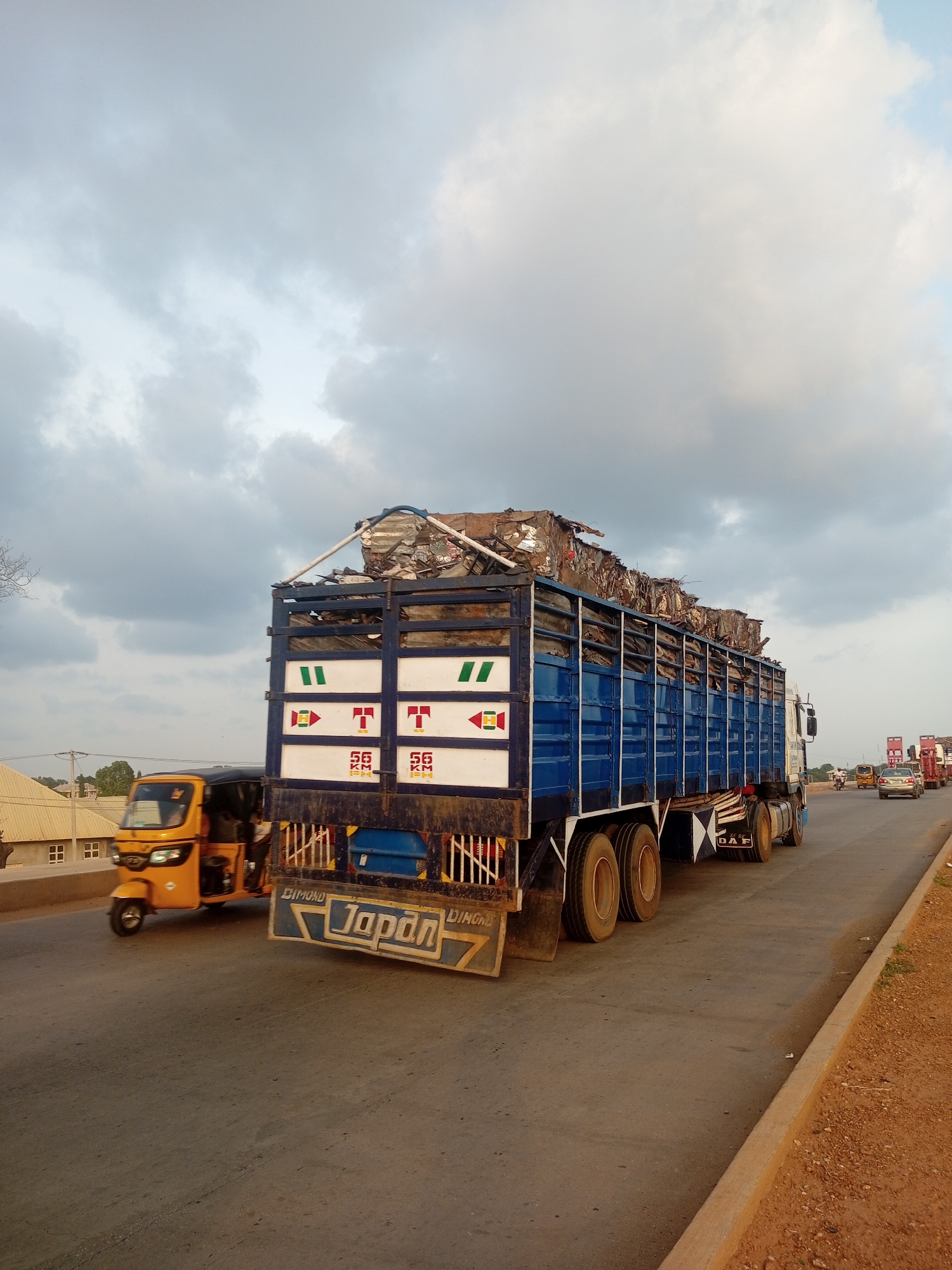 A large, blue flatbed truck is driving on a road. The truck is loaded with a significant amount of scrap metal, which is piled high above the truck's bed. The truck has white panels on its sides with red and green markings. There is visible text on the truck that reads "DIMOND JAPAN".
To the left of the truck, a yellow three-wheeled vehicle, often referred to as an auto-rickshaw or tuk-tuk, is also moving on the road. In the background, there are buildings with sloped roofs and trees. The sky is filled with dramatic, cloudy formations, suggesting it could be late afternoon or early evening, or that the weather is changing. The road appears to be a multi-lane highway, with other vehicles visible further down the road, including a silver car. The scene depicts a transportation of goods, specifically scrap metal, on a public road.