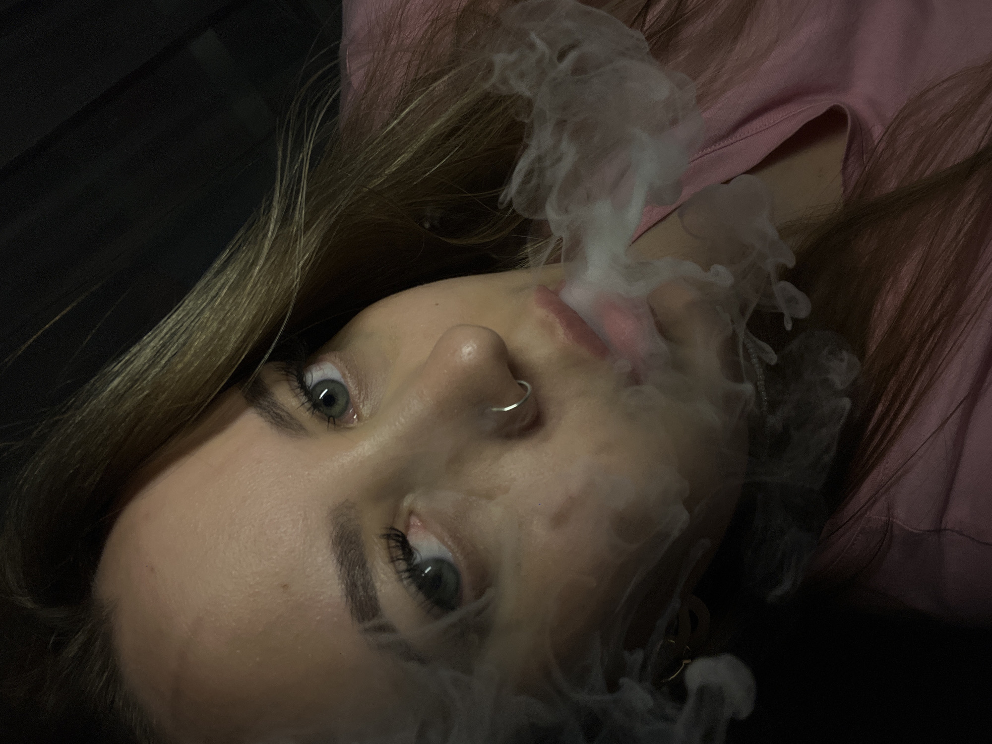 A young woman with blonde hair and gray eyes is exhaling smoke. She has a nose ring in her left nostril and appears to be wearing a pink top. The background is dark. There is no information to suggest the image was taken in the city of Novopokrovskaya, Russia.