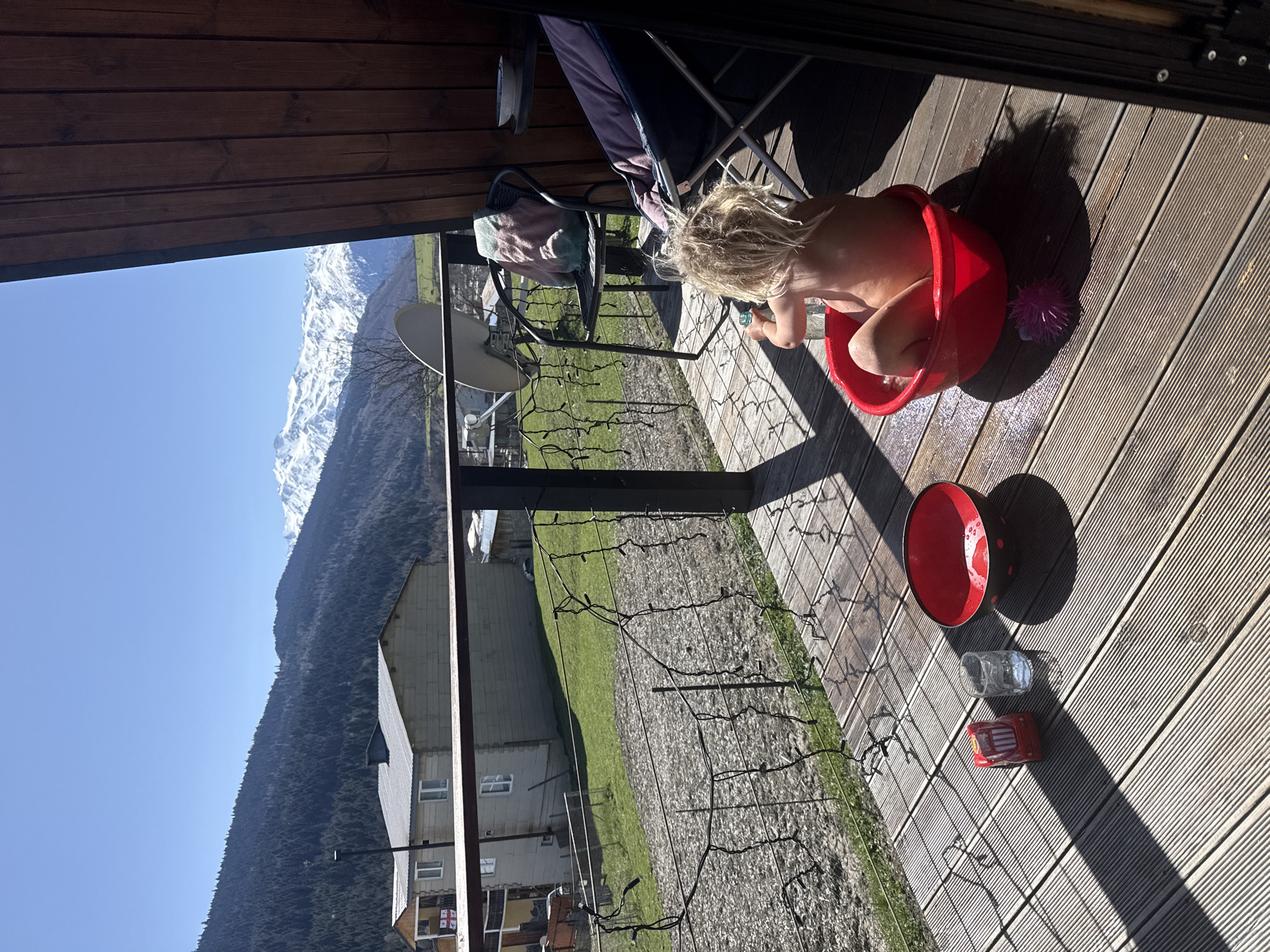 The image depicts a bright, sunny scene on a wooden deck or balcony, overlooking a mountainous landscape. The central subject is a young, fair-haired child, appearing partially nude, seated inside a red plastic bucket filled with water, engaged in playful activity.
On the wooden deck, next to the child's bucket, are a purple spiky toy, a clear glass, and a small red toy car. Another empty red bowl or lid is also present on the deck. The strong shadows cast by objects and the deck railing indicate bright sunlight, suggesting it is late morning or early afternoon. Above, a wooden ceiling or overhang is visible.
To the left, a dark-colored folding chair with a purple cushion and a light-colored towel draped over it can be seen. A satellite dish is mounted on the railing, which is also adorned with decorative string lights. Beyond the deck, a rural scene unfolds with green grassy areas, interspersed with a few buildings and houses. One of the houses in the distance clearly displays a small Georgian flag on its facade. The background is dominated by majestic, snow-capped mountains under a clear blue sky, with a forested mountain range also prominent. The overall atmosphere is peaceful and playful, capturing a moment of outdoor enjoyment in a picturesque mountain setting in Mestia, Georgia.