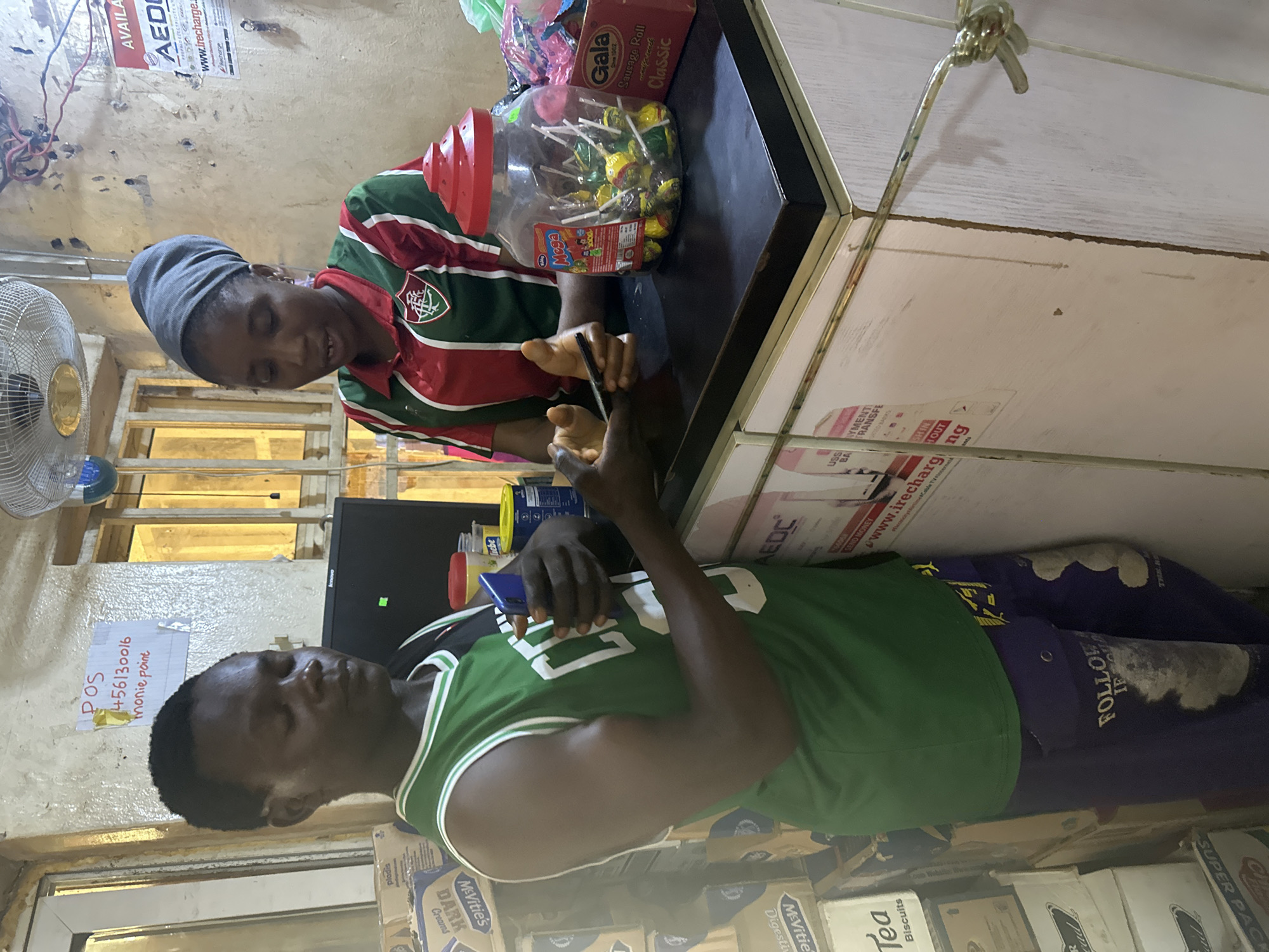 In a cluttered convenience store in Abuja, Nigeria, a man in a green sleeveless jersey and purple patterned pants holds a blue smartphone. He is interacting with a woman wearing a grey headscarf and a red, green, and white striped shirt. She is behind a counter, which holds a large jar of lollipops, a red box of "Gala Sausage Roll Classic," and various other products. The background features shelves stacked with goods, a computer monitor, and advertisements on the wall, including one for AEDC (Abuja Electricity Distribution Company). The overall impression is of a busy, everyday transaction in a local retail setting.