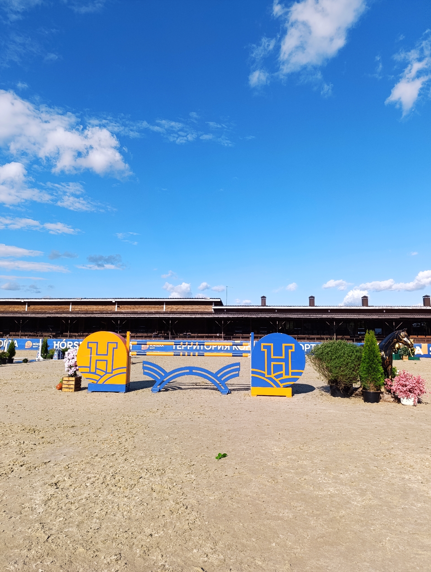 This media file depicts an outdoor equestrian event, likely a show jumping competition, taking place on a sandy arena in Ivanovskoe, Russia. The scene is set under a bright blue sky with scattered white clouds, suggesting a clear and pleasant day. The time of day appears to be daytime, possibly late morning or early afternoon, given the lighting and shadows.
The main subjects in the foreground are two large, decorative jump elements. The jump on the left is a circular panel with a prominent yellow "H" logo against a blue background, featuring stylized yellow waves at the bottom. It is placed next to a wooden planter filled with pink flowers and a small orange pumpkin. To the right of this is a blue, arched jump element. The jump on the far right is another circular panel with a similar yellow "H" logo, also against a blue background, and next to it is a black, stylized horse sculpture. Green bushes and potted plants are strategically placed around the jumps, adding to the visual appeal of the competition grounds.
In the background, a long, wooden stable or grandstand building with a brown roof and visible chimneys dominates the scene. The building has large open doorways and windows, suggesting it houses horses or spectators. Various banners and signs are visible along the front of the building and on the arena's perimeter fence. One prominent banner displays the text "HORS", and another clearly shows the Cyrillic text "ТЕРРИТОРИЯ" (Territory) along with logos that include "HORS SPORT". These banners indicate sponsorship or the name of the event or venue.
The overall impression is one of a well-organized and picturesque equestrian event, with attention to detail in the decorations and the presentation of the venue. The sandy arena is well-maintained, and the clear sky contributes to a positive and inviting atmosphere. No people are clearly visible in the image, suggesting the focus is on the course itself and the decorative elements.