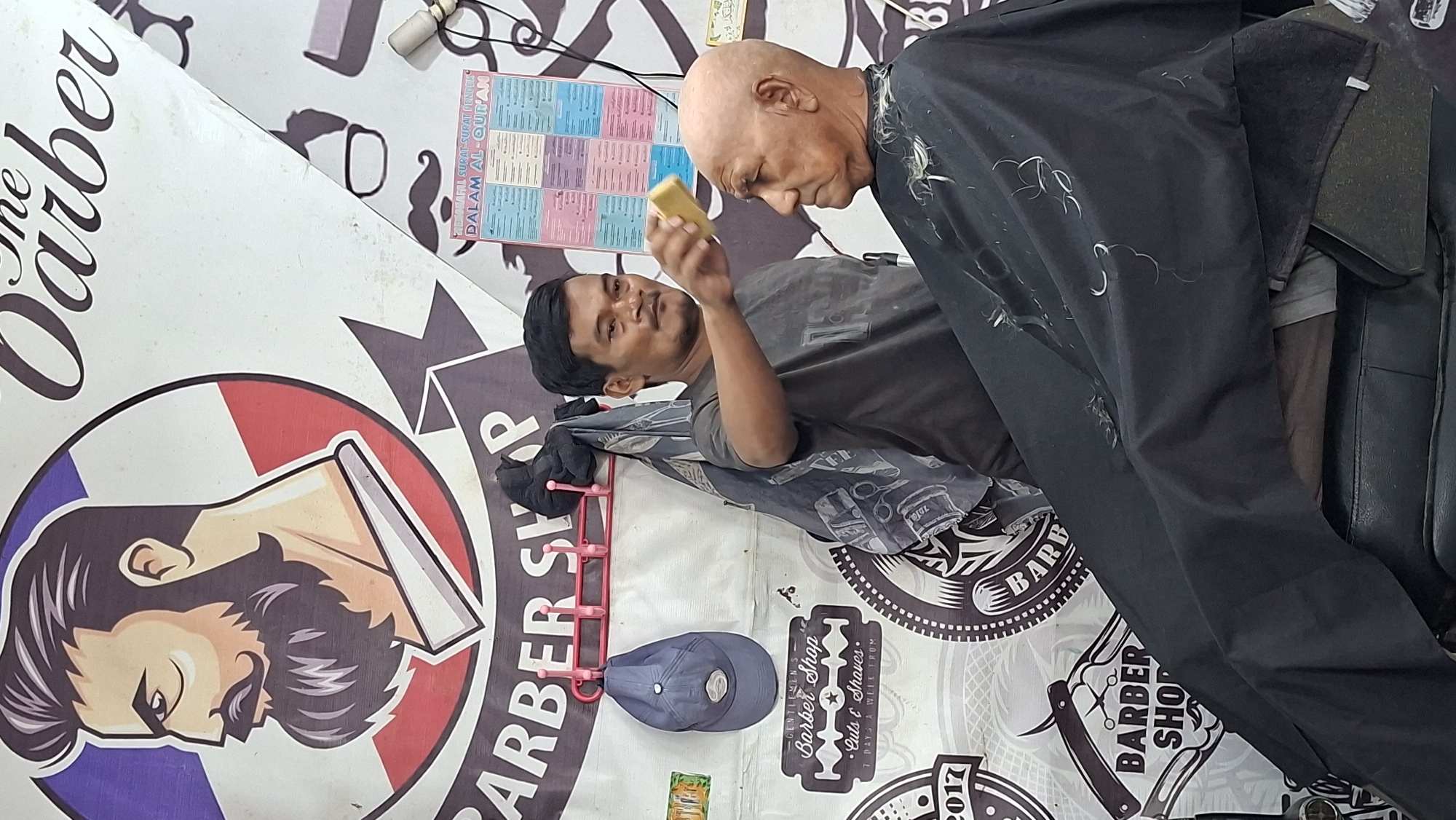 This is a medium shot, eye-level, indoor photograph taken at a barber shop. The barber shop is decorated with various Barber Shop logos and a poster with text in Arabic and Indonesian. The overall lighting is bright, coming from an unseen source. The photograph focuses on a barber, an adult male with dark hair, who is holding a yellow sponge in his right hand and is in the process of shaving the head of an elderly man. The elderly man is bald and has a serious expression on his face, and he is wearing a black cape that covers his body. The barber shop appears to be a local establishment, with the Barber Shop logos and posters suggesting it is a business that caters to the local community.