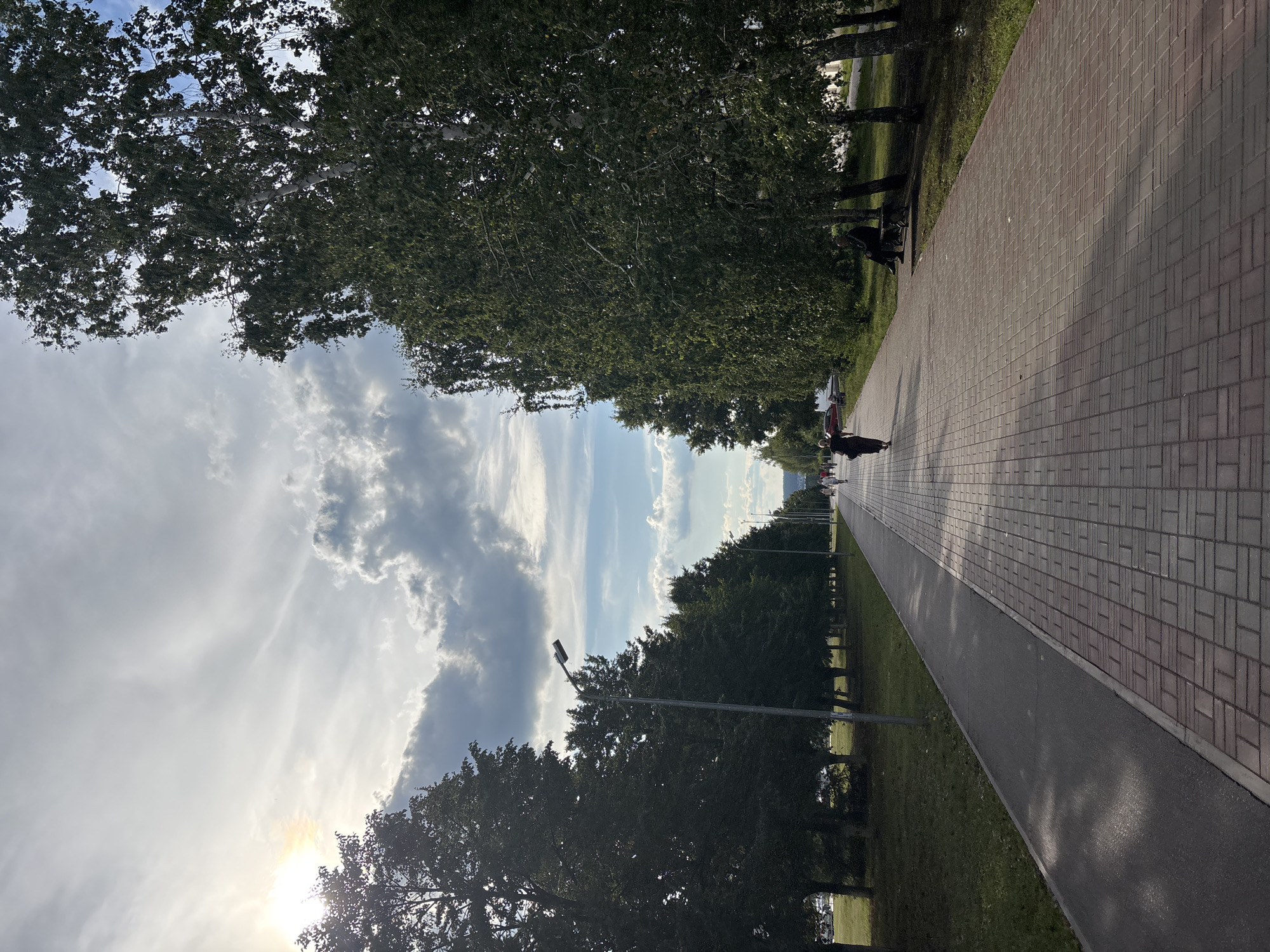 The image depicts a paved walkway lined with trees and grass, leading into the distance. The walkway is made of red brick pavers and has a separate asphalt path running alongside it. Several people are walking along the path, and one person is sitting on a bench under a tree. The sky is partly cloudy with the sun shining through, creating shadows on the walkway. Streetlights are visible along the path. The scene appears to be in a park or public area in Naberezhnye Chelny, Russia.