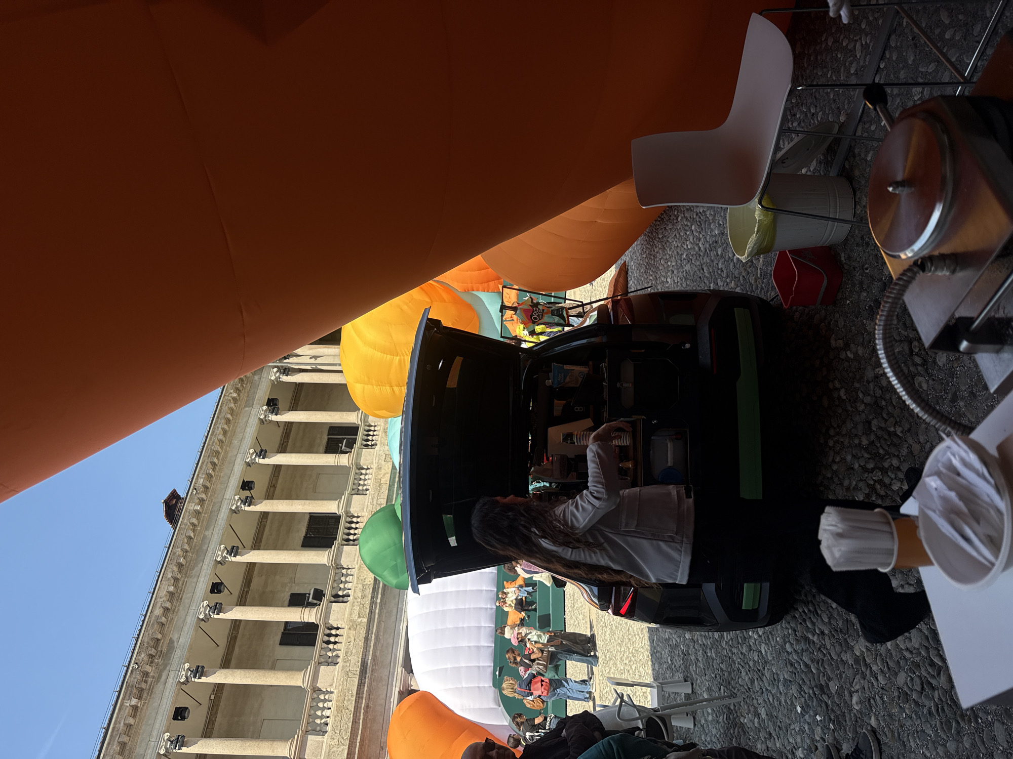This image captures a vibrant outdoor event in Milan, Italy, under a clear blue sky during the daytime. The scene is set in a historic courtyard, with a grand classical building featuring multiple columns, balconies, and an ornate facade visible on the left side of the frame. The ground is paved with cobblestones.
In the center, a dark-colored mobile food or drink vendor vehicle has its rear hatch open, revealing equipment inside. A woman with long, dark, wavy hair, wearing a white jacket, is actively working within the vehicle, seemingly preparing beverages or food. To the right of the vendor, partially obscured by an orange inflatable, are a white chair, a white bucket with a yellow liner, a red bucket, and a metallic food preparation machine. A stand holding disposable cups and what appear to be stirrers or packets is on a surface next to the vendor area.
Large, colorful inflatable structures are prominent throughout the scene. A substantial orange inflatable dominates the right foreground, curving upwards. Other yellow, green, and white inflatable structures are visible in the background, drawing attention. A crowd of people, mostly adults, are gathered in the middle ground between the vendor and the classical building, observing or participating in the event. They are clustered around the various inflatables. The overall atmosphere appears lively and relaxed, characteristic of a public festival or fair. No discernible text is visible in the image.