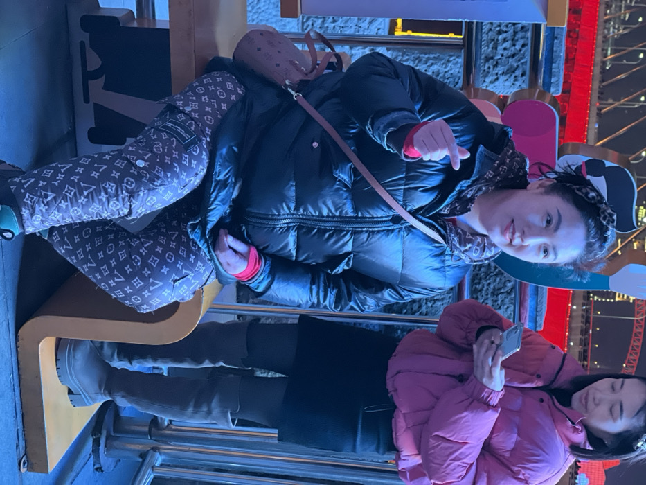 An outdoor nighttime scene in Yuzhong District, China, depicts two East Asian females illuminated by artificial blue light, with distant red urban lighting. A woman, appearing middle-aged, is seated on a yellow, minimalist bench-like structure. She wears a black puffer jacket over a patterned hoodie and a red long-sleeved shirt, brown pants with an "LV" monogram pattern, and a leopard print headband. A brown "MCM" monogrammed cross-body bag is visible. She makes a finger heart gesture with her right hand, her left hand out of view, and smiles faintly. To her right, a second woman stands, holding a silver smartphone with both hands and looking at the screen. She wears a pink puffer jacket, a black mini skirt, and light brown over-the-knee boots. A dark, textured wall or barrier is behind them, and a metallic railing is present. In the far background, a bridge or large structure is distinctly illuminated in red.