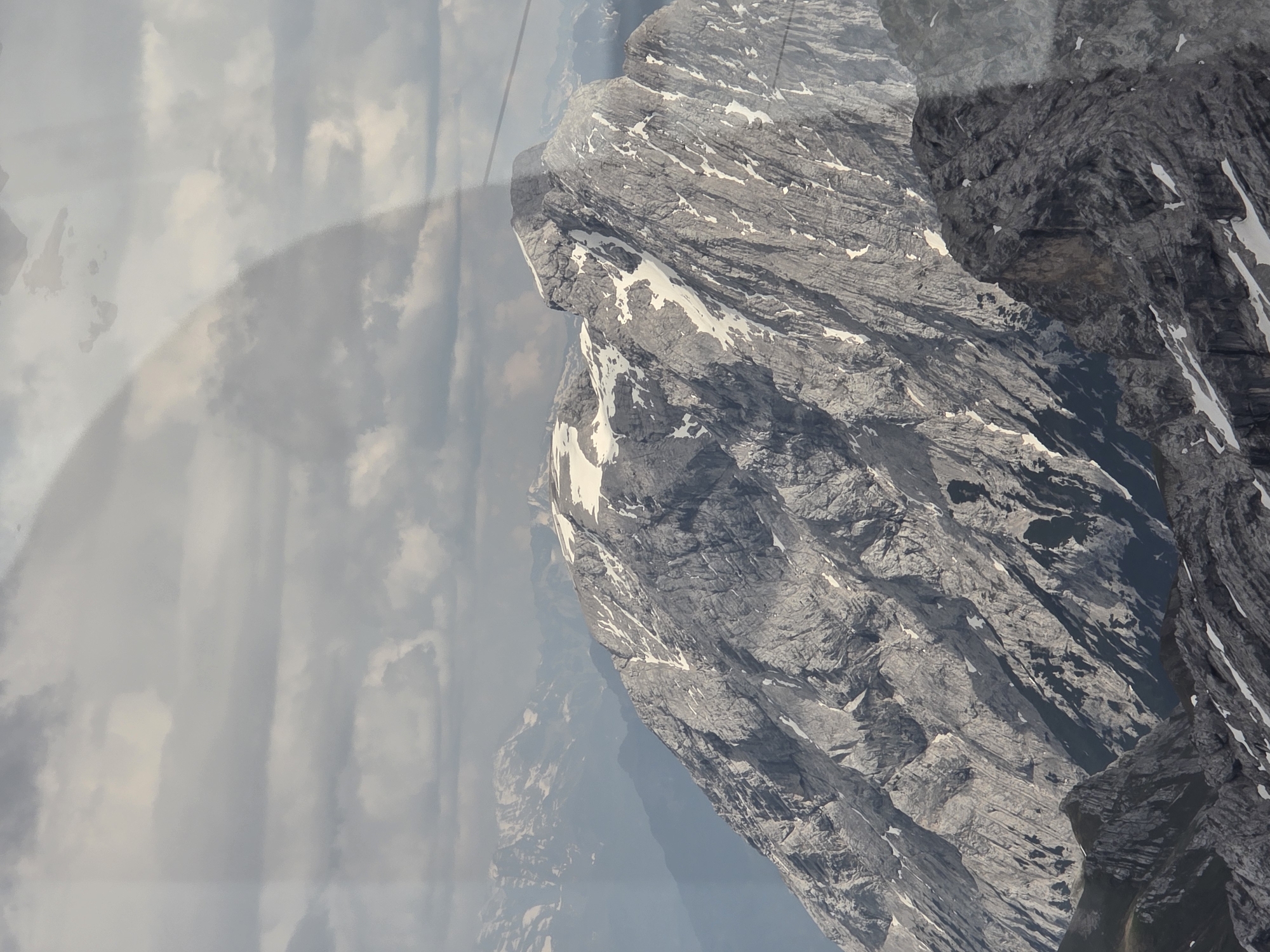 An overhead, wide shot captures a majestic, jagged mountain range under a cloudy sky. The mountains are predominantly gray, with patches of white snow clinging to their rocky slopes. The sharp, uneven terrain suggests a high-altitude environment, possibly with a cable car or observation point providing this elevated perspective. The sky is filled with dramatic, overcast clouds, with lighter patches hinting at sunlight breaking through. A faint shadow, possibly from a structure or a person, is visible across the upper portion of the image. The overall scene conveys a sense of vastness and the raw beauty of nature.