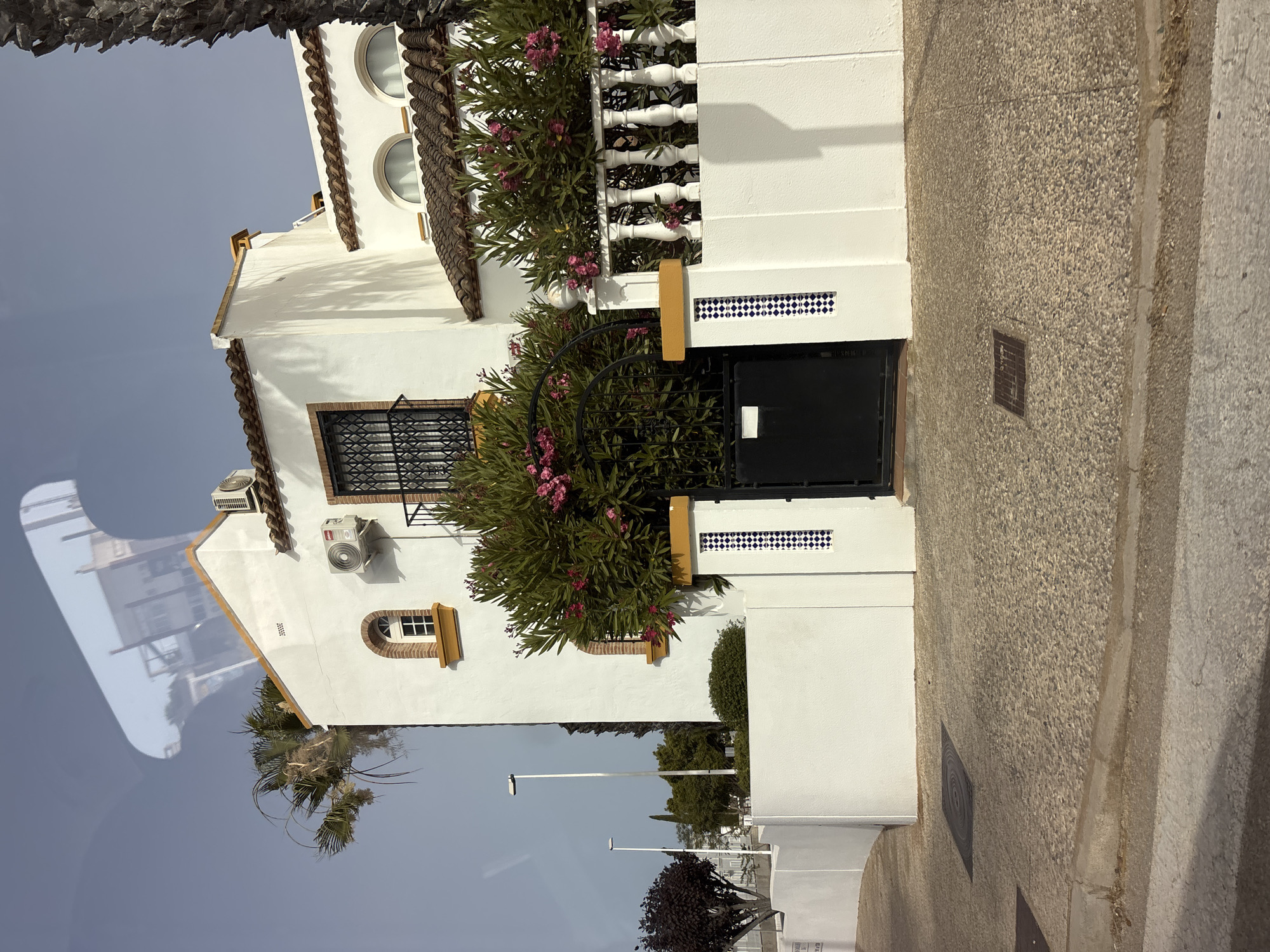 The image shows the exterior of a white, multi-story building with red tile roofing, typical of Mediterranean architecture. The building features arched windows with decorative yellow trim, and a black wrought-iron security gate and balcony railing. It is surrounded by a white wall, with a paved driveway and sidewalk in the foreground. A dense bush with pink flowers is planted along the wall, bordering the entrance. In the background, there are streetlights and some trees, including a palm tree. The sky is clear and blue. The scene appears to be in a residential area within the city of Orihuela, Spain, given the architectural style and climate. No people are visible in the image, and there are no apparent actions or interactions taking place. The overall impression is one of a well-maintained private residence.