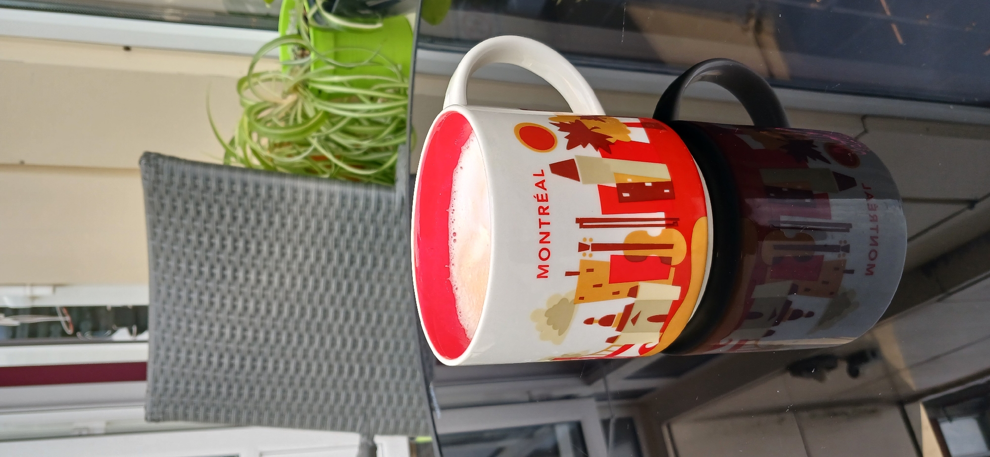 This image shows a close-up of two mugs on a reflective surface, likely a table. The mug in the foreground is white with a red rim and features a design of the Montreal skyline, complete with maple leaves and buildings. The text "MONTREAL" is clearly visible on the side of this mug. The second mug, dark in color, is partially obscured by the first and its design is less discernible, though it appears to have a similar skyline graphic reflected on its surface.
The mugs contain a light-colored liquid, possibly coffee or tea, with some foam on top. In the background, to the left, a potted plant with long, striped green leaves is visible. A woven gray chair is also present in the background, suggesting an outdoor or patio setting. The overall lighting suggests it is daytime. The reflective surface of the table shows reflections of the mugs and the plant. The scene is set in Khmelnytskyi, Ukraine.