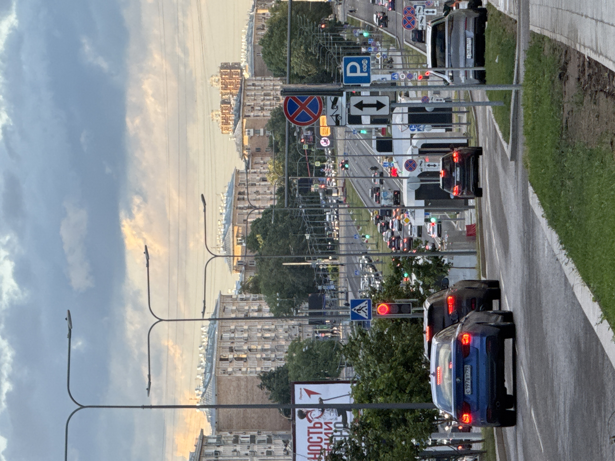 The image shows a street scene in a city with tall buildings in the background. Several cars are on the road, and there are traffic lights and signs. The sky is cloudy with some hints of sunlight. On the left side of the image, there is a large billboard with text in Russian. The foreground shows a grassy area and a sidewalk. The road appears to be a multi-lane street with traffic moving in both directions. There are streetlights visible along the road.