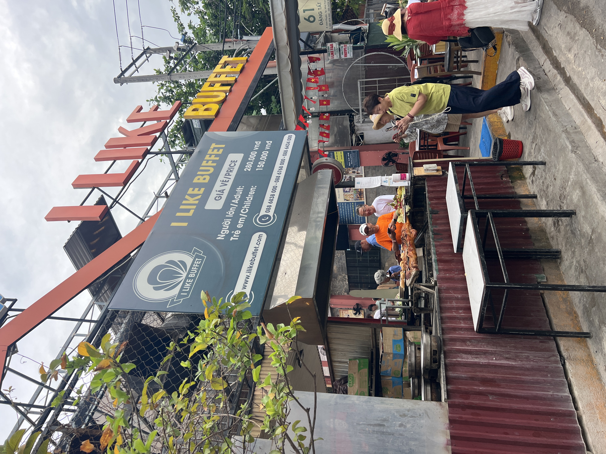 The image depicts the exterior of "I LIKE BUFFET" restaurant located at 61 Đặng Tất Street in Vĩnh Phước Ward, Nha Trang city, Vietnam. The restaurant has a prominent sign with its name in large, red lettering above smaller, yellow "BUFFET" lettering. A secondary sign advertises the prices, 280,000 VND for adults and 150,000 VND for children, and includes the website and phone numbers.
People are visible at the restaurant. One person is looking at their phone, another is wearing a large hat and red shirt. Two people are working at the buffet.
The restaurant exterior includes Vietnamese flags, tables with white tops, and a corrugated metal facade. There are various stacked boxes and equipment near the buffet area. Power lines and trees are visible in the background.