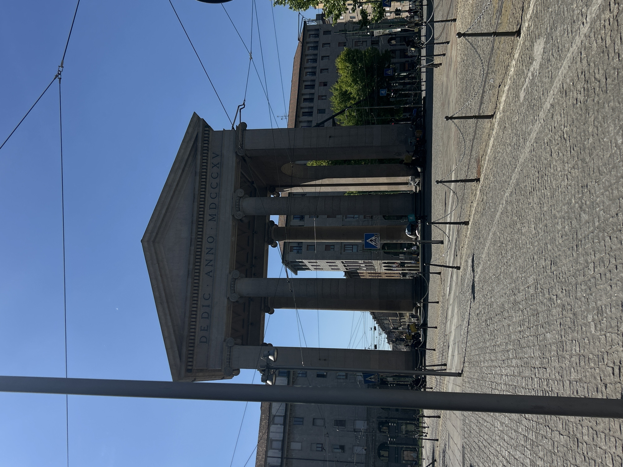 This is a medium, eye-level shot taken outdoors in Milan, Italy. The image features a stone triumphal arch structure in the center, with inscription "DEDIC . ANNO . MDCCCXXV" on the front, denoting the year 1825 in Roman numerals. The archway consists of large pillars supporting a triangular pediment. The structure sits on a cobbled street. Beyond the archway, buildings are visible. Street furniture, including poles and chains, lines the edge of the street. Overhead tram lines are visible. The sky is a clear, light blue.