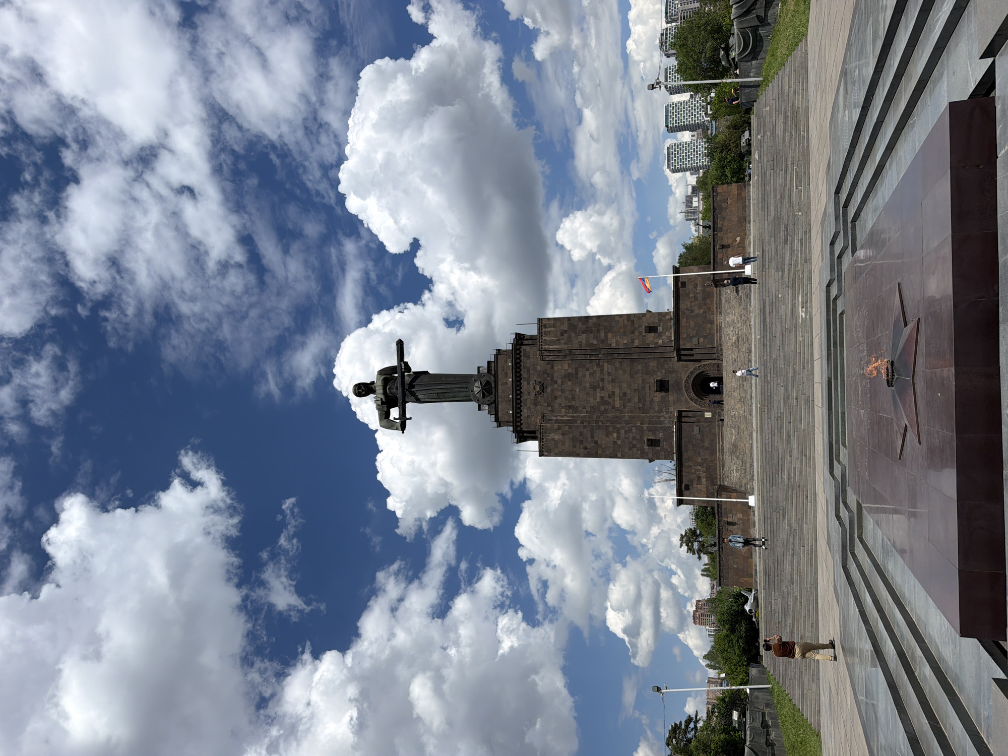 The image shows the Mother Armenia statue and Eternal Flame at Victory Park in Yerevan, Armenia, set against a backdrop of a blue sky with numerous white cumulus clouds. The statue, rendered in dark metal, stands atop a tall, brown stone structure. Below it, a series of stone steps lead towards a large, reddish-brown monument featuring a star shape at its center, where an eternal flame burns. Several people are visible on the steps, including individuals walking and taking photographs. A small Armenian flag is visible on a pole near the statue's base. Beyond the park's vegetation, high-rise buildings can be seen on the horizon. The photograph appears to be taken from a low angle, emphasizing the height of the statue and monument.