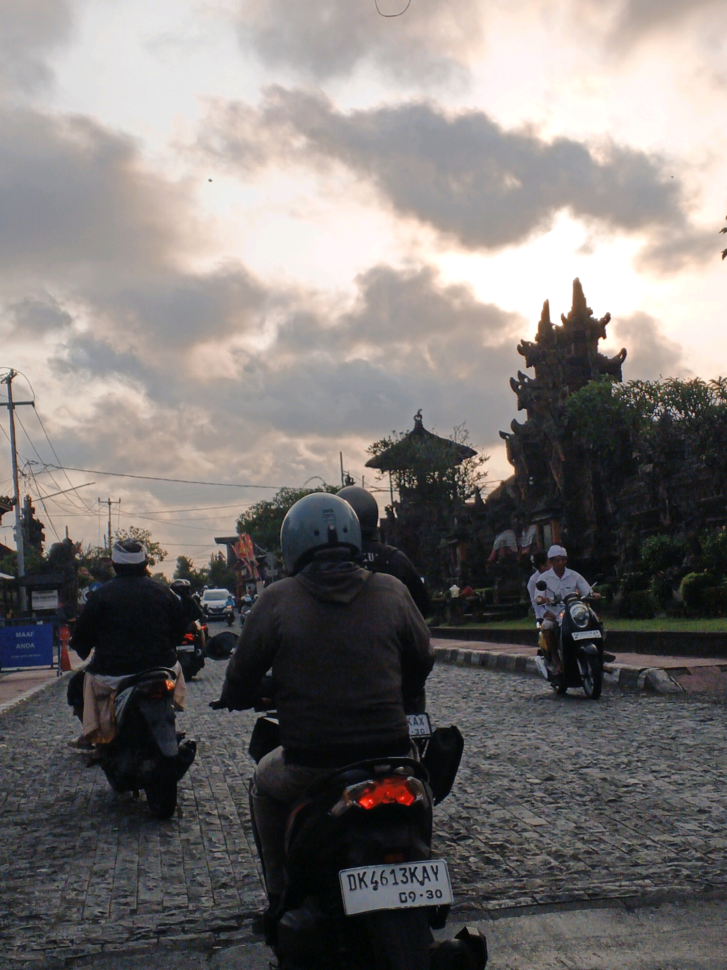 In Sukawati, Indonesia, a group of people are riding motorcycles on a cobblestone street. The sky is cloudy with the sun setting behind a traditional Balinese temple. Two men are riding motorcycles in the foreground, their backs to the camera. One is wearing a grey helmet and jacket, while the other is wearing a black jacket and a white headscarf. A third motorcycle is visible in the middle ground, ridden by a man in a white shirt and a white headscarf. The temple in the background is ornately decorated with carvings and lush greenery. Power lines are visible in the upper left corner of the image, indicating an urban or semi-urban setting.