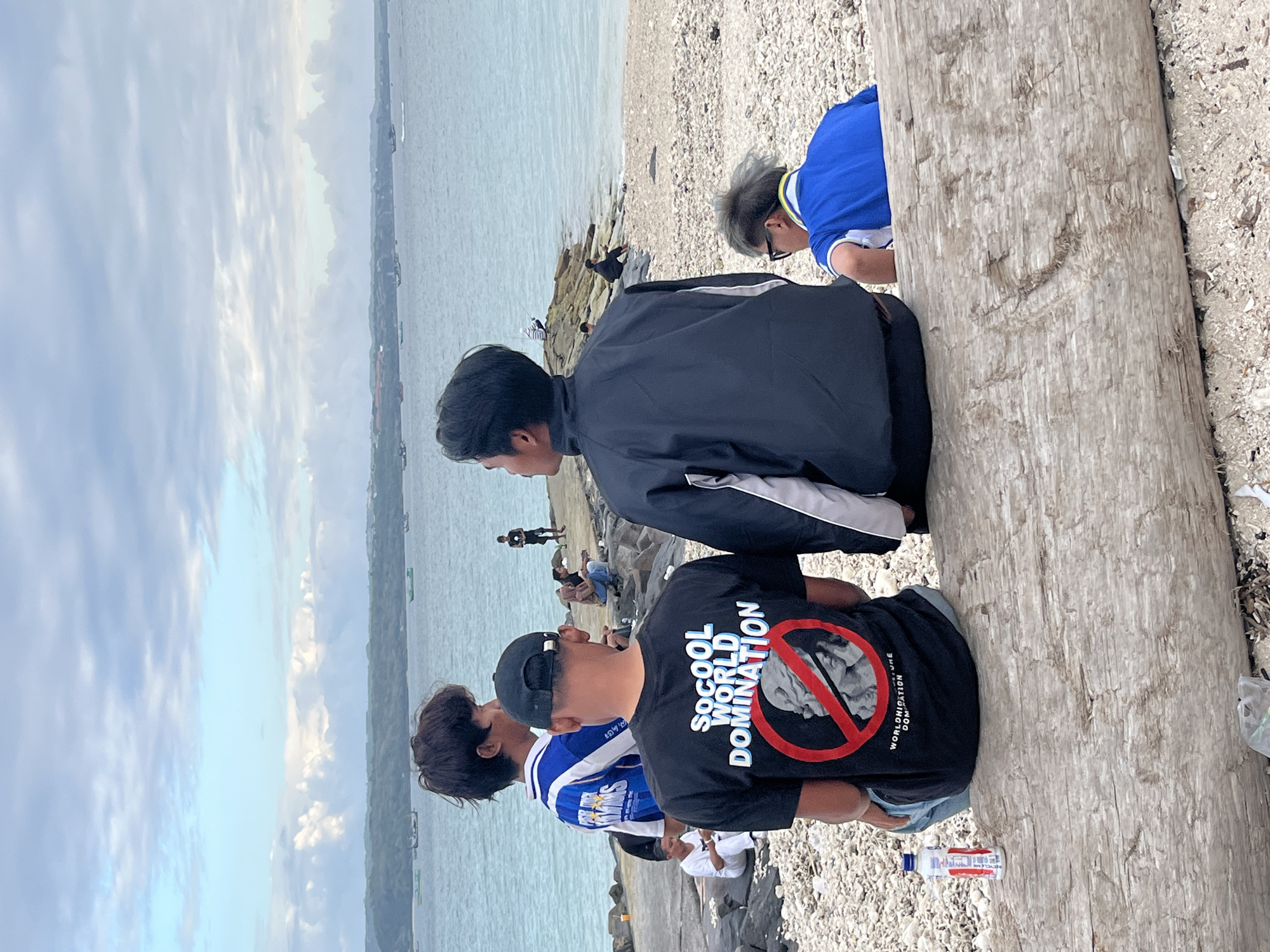 In Kuta, Indonesia, a group of young men are gathered on a rocky beach near the ocean. The sky is overcast with clouds, and several boats are visible on the water.
In the foreground, four young men are seated on a large piece of driftwood. One man wears a black t-shirt with a graphic that reads "SOCOOL WORLD DOMINATION" and a red circle with a line through it, superimposed over a statue's head. Another man is wearing a black jacket with white accents. A third man wears a blue and white striped shirt. The fourth man is partially visible and appears to be wearing a blue polo shirt. A plastic bottle of water is placed on the driftwood.
In the middle ground, other individuals are scattered across the beach. One person is sitting on the rocks and looking out at the water. Another person, wearing a white shirt and pants, is seated on the rocks and looking down. Further in the distance, on a rocky outcrop, a person is standing and looking towards the sea, possibly fishing.
The environment is a beach with a mixture of sand and pebbles. The ocean is visible in the background, and a distant coastline with greenery and buildings can be seen. The overall scene depicts a casual gathering of young people by the sea.