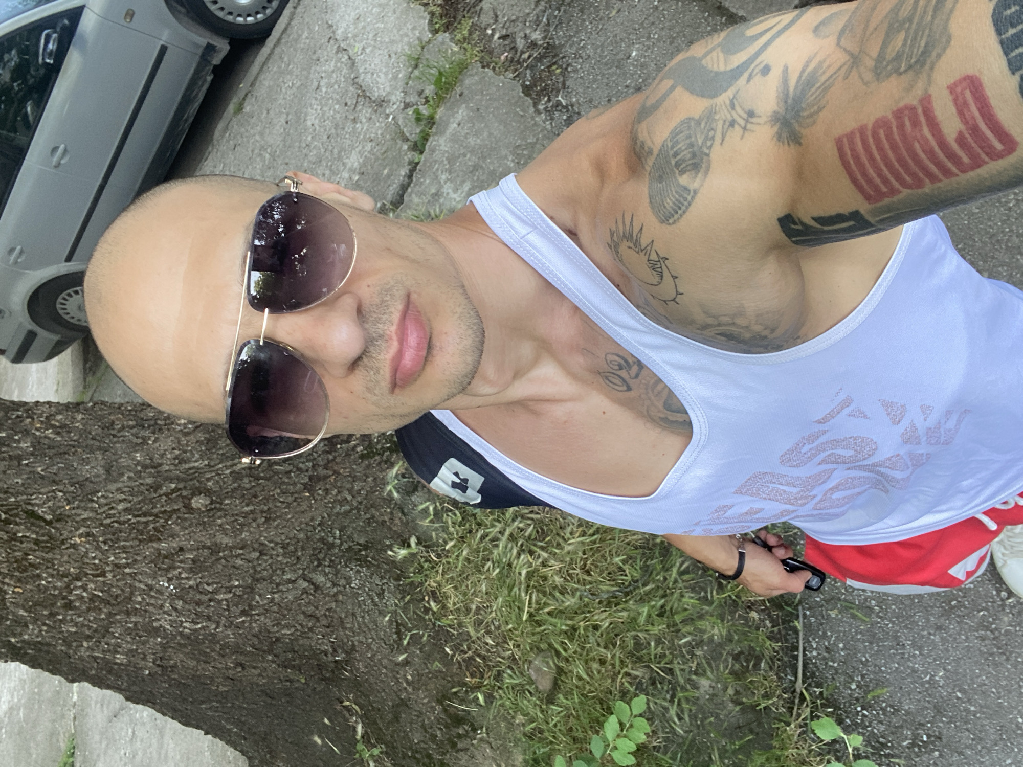 A man with a shaved head and sunglasses poses for a selfie on a sidewalk in Pleven, Bulgaria. He is wearing a white tank top, red shorts, and white sneakers. He has tattoos on his arms and chest. In the background, a tree, grass, and a silver car are visible. The man is holding a set of keys in his right hand.