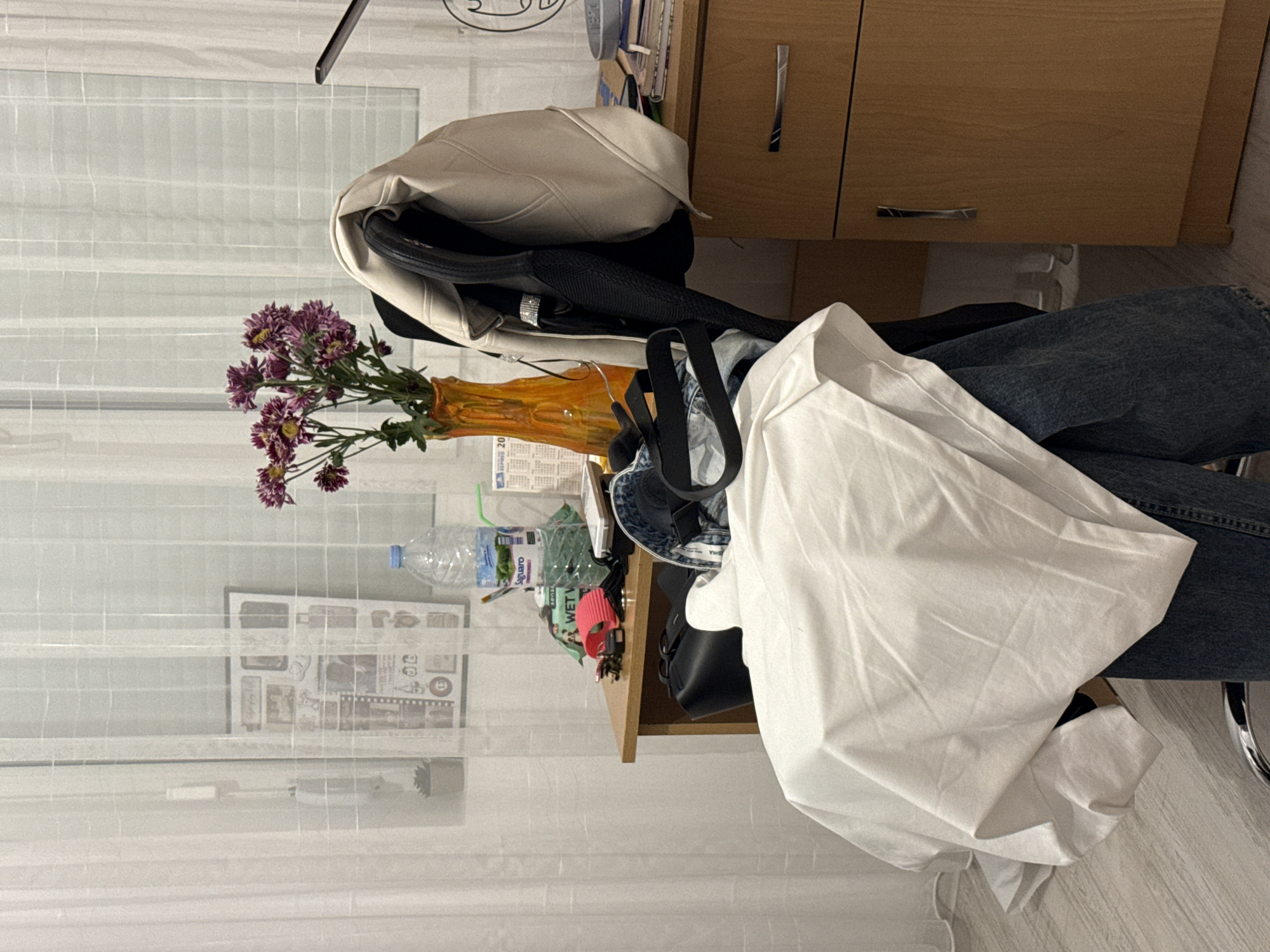The image shows a cluttered desk in a room with white, semi-transparent curtains. A bouquet of purple flowers in an orange vase sits on the desk near a water bottle, various papers, and a red hair tie. Several handbags and a beige jacket are piled on the desk. A portion of a wooden cabinet is visible in the upper right corner, and what appears to be a pair of jeans is draped over the chair. The location is specified as Blagoevgrad, Bulgaria, although there are no discernible landmarks to confirm this.