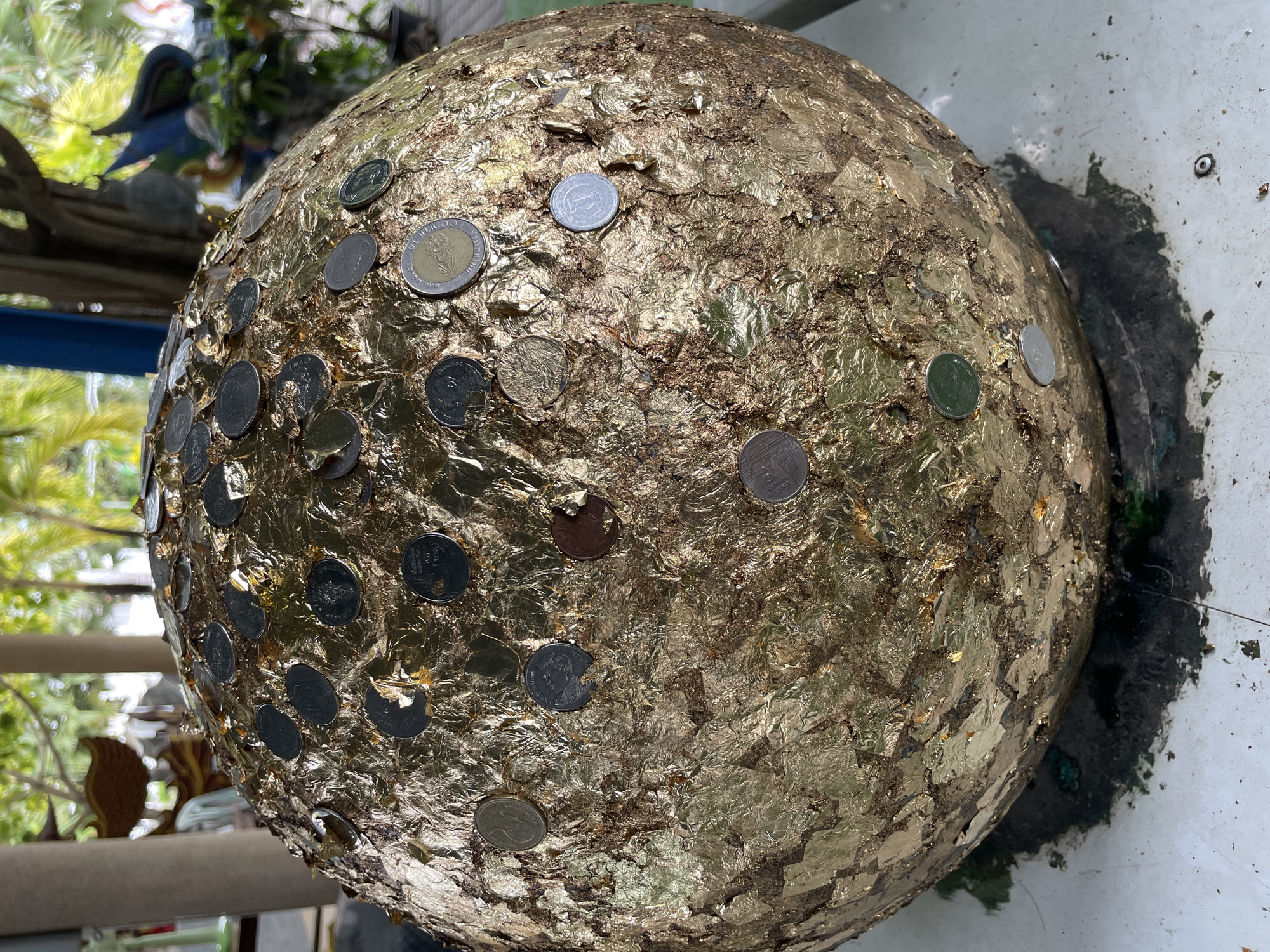 In Tambon Na Chom Thian, Thailand, a large, spherical object, possibly a stone or sculpture, is covered in gold leaf and adorned with numerous coins. The object rests on a white surface, with a dark, wet area visible beneath it, suggesting it might be in an outdoor or partially exposed location. In the background, blurred greenery and trees indicate an outdoor setting, possibly within temple grounds or a garden. The coins, a mix of silver and gold-toned, are pressed into the gold leaf, creating a textured and visually striking surface. The lighting suggests it is daytime, with natural light illuminating the scene. No people are visible in the frame.