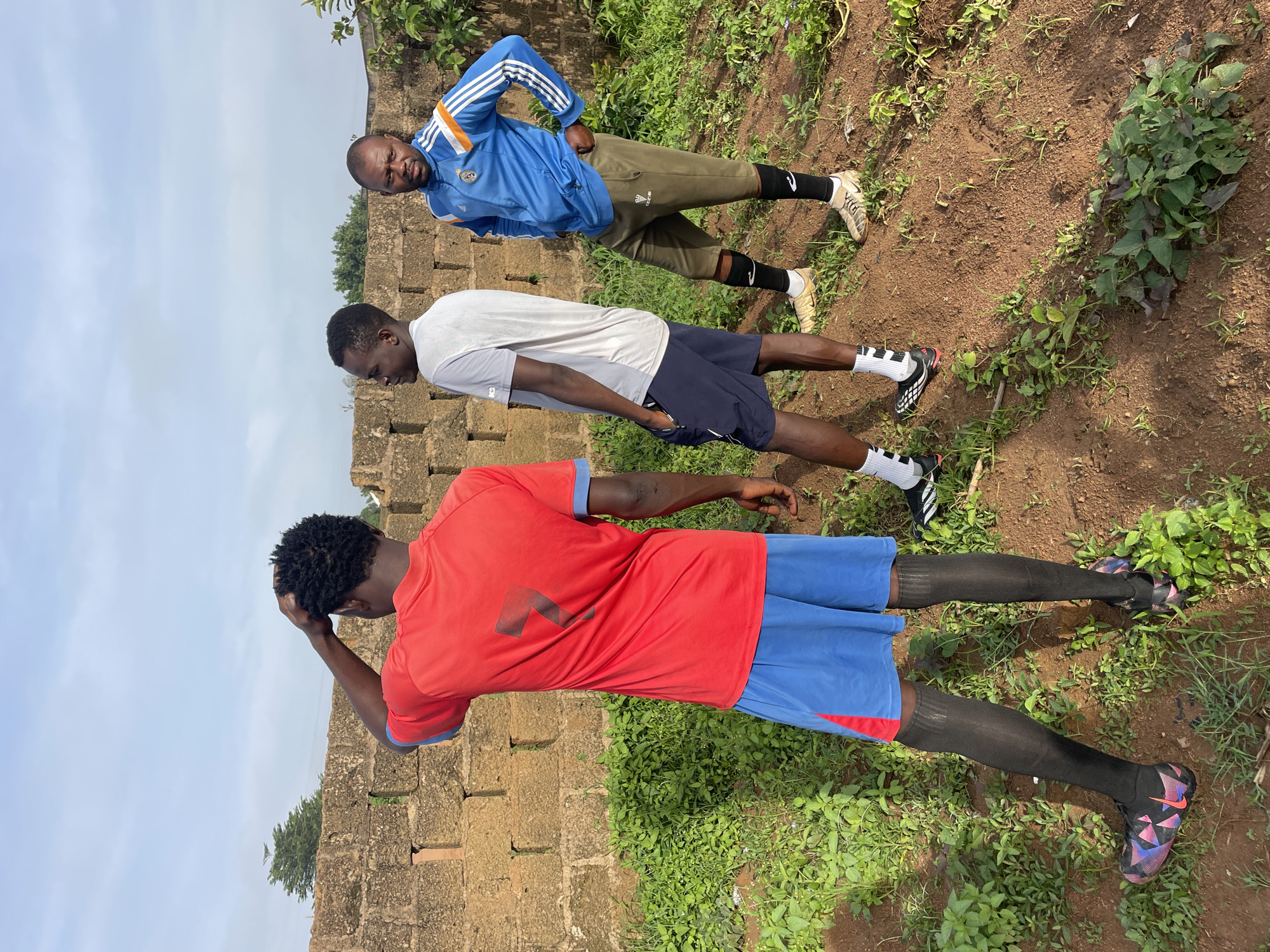Three individuals are standing in an outdoor setting, likely in Gura Top, Nigeria, with a brick wall in the background. The person in the foreground, on the left, is a Black male wearing a red t-shirt with the number "7" in black on the back, and blue shorts with red accents. He is also wearing black socks that extend to his knees and colorful soccer cleats. His right arm is raised, with his hand on the back of his head.
To his right stands another Black male in a light gray t-shirt and navy blue shorts. He is looking to his left, with his hands in his pockets.
The third Black male is on the far right, wearing a blue Adidas track jacket with white and orange stripes on the sleeves, and olive green capri-length pants. He has on black socks and light-colored soccer cleats. His left hand is on his hip, and he is looking towards the viewer with a neutral expression.
The ground is composed of reddish-brown soil with patches of green grass and some leafy plants. The brick wall behind them is weathered and appears to be constructed from rough-hewn, reddish-brown bricks. The sky above is overcast with some visible clouds. The overall impression is of a casual, possibly sports-related gathering in a rural or semi-urban environment.