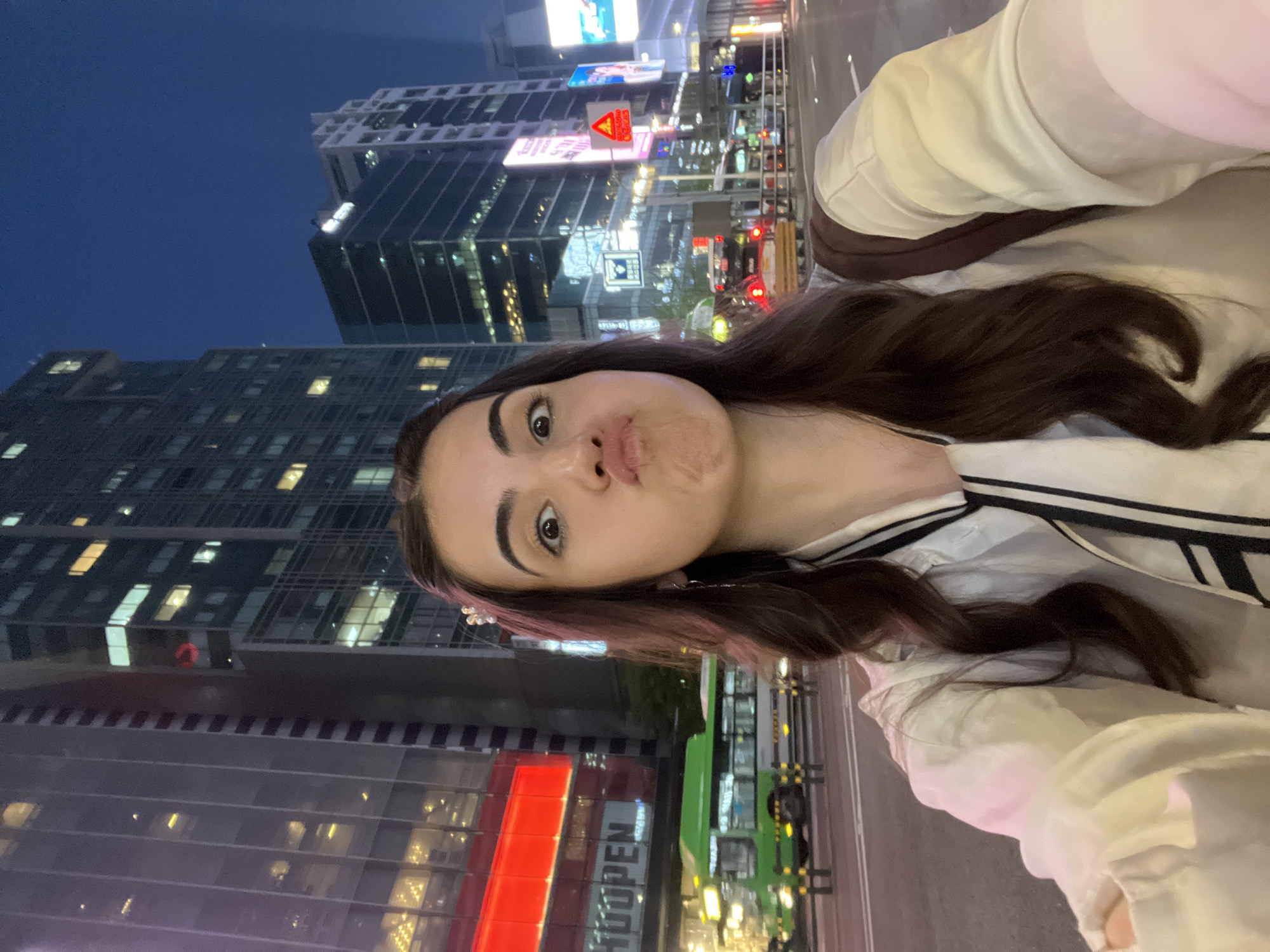 A young woman takes a selfie in Seoul, South Korea at night, with the bustling city skyline behind her. The woman has dark hair with pink highlights and is wearing a white shirt with black stripes. She is making a kissing face towards the camera. In the background, tall skyscrapers are visible, illuminated with lights. Streetlights and traffic signals add to the vibrant urban atmosphere. Some buildings display prominent signs like "HUOPEN".