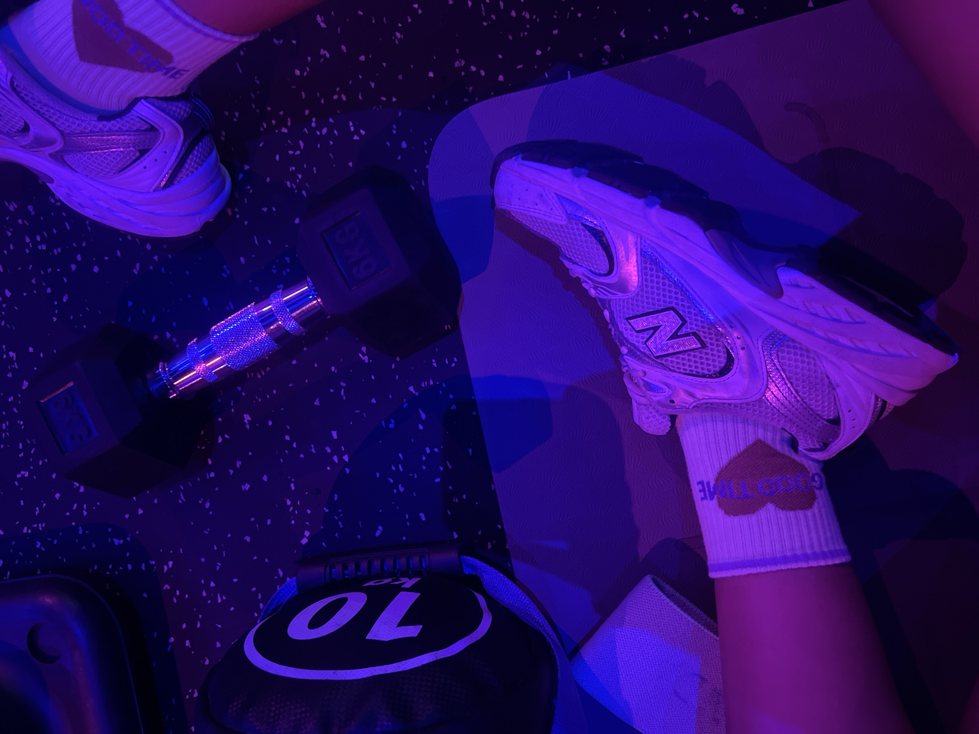 The image captures an overhead view of an indoor gym or exercise setting, characterized by striking purple and blue lighting that creates a vibrant, almost neon atmosphere. The scene is dominated by a person's legs and feet, wearing athletic shoes and socks, surrounded by fitness equipment on a dark, speckled gym floor.
The person wears white athletic socks. On the visible right sock, the text "GOOD TIME" is emblazoned within a heart shape, rendered in a light brown or peach color. A similar design is partially visible on the left sock. Both feet are clad in stylish sneakers featuring mesh and metallic silver accents, one of which displays a prominent "N" logo on its side.
Various exercise tools are present. A silver dumbbell with hexagonal ends is prominently placed on the floor, clearly marked with "6KG." Beneath the left foot, a dark, textured exercise or yoga mat is visible. In the bottom left portion of the image, a black weight bag or sandbag is partially seen, featuring a large white number "10" and the fragmented letters "KG." Another light-colored elastic resistance band or strap lies near the right foot, resting on the mat.
The overall impression is a pause during a workout session, set in a modern gym environment with a distinct, dynamic lighting scheme.