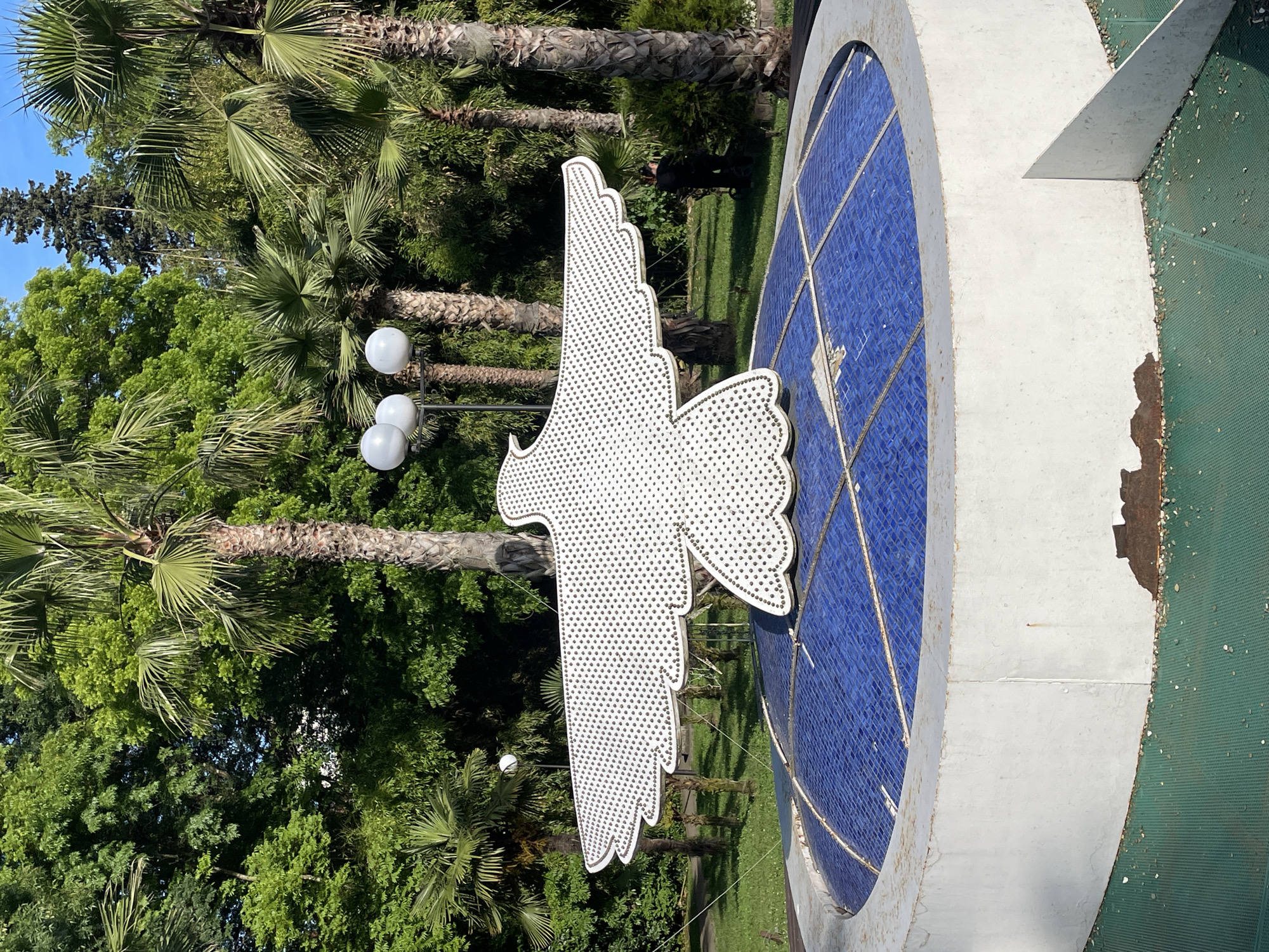 An outdoor shot captures a white, stylized bird sculpture with outstretched wings positioned above a circular fountain in Sochi, Russia. The bird sculpture is adorned with numerous small, dark dots, creating a textured surface. The fountain features a domed top covered in blue tiles, with a white concrete base. The surrounding environment is lush with greenery, including palm trees and dense foliage. In the background, a lamppost with three spherical lights is visible, and a person dressed in dark clothing is seen in the middle distance, appearing to be tending to the plants. The scene is illuminated by natural daylight, suggesting it is daytime.