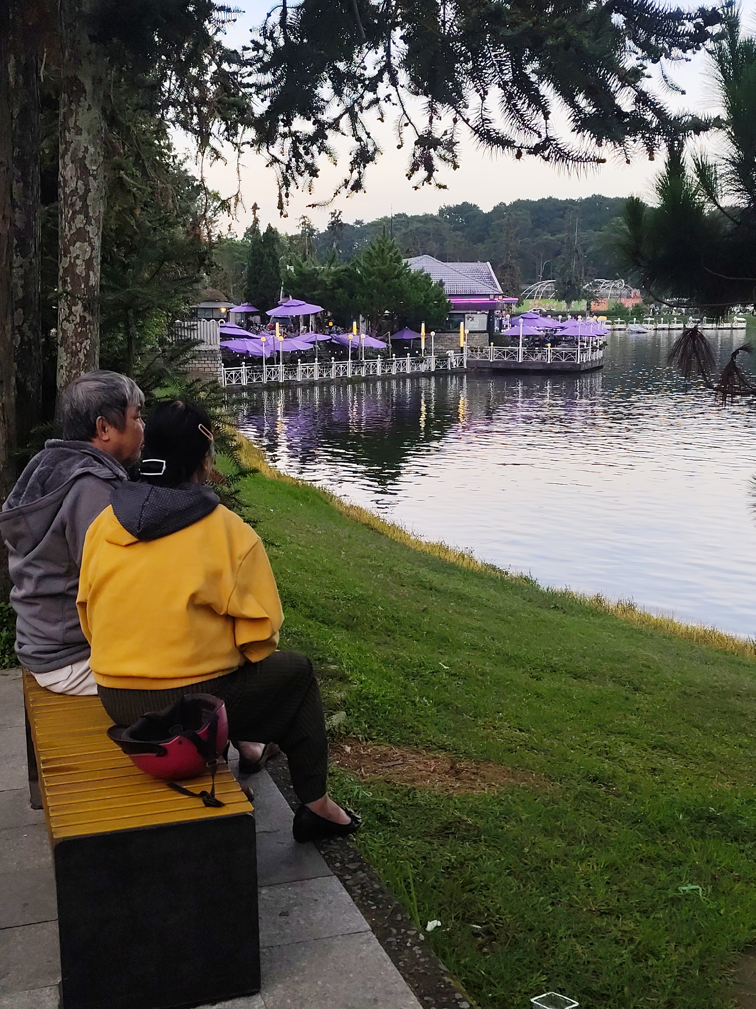 In Da Lat, Vietnam, an elderly couple is seated on a bench facing a calm lake. The woman, wearing a yellow hoodie and dark pants, has her back to the viewer, while the man, in a grey hoodie and white shirt, is seated to her left, also facing the lake. A pink helmet rests on the bench between them. The background features a restaurant with purple umbrellas and seating areas on the water, surrounded by trees. The lake reflects the twilight sky and the lights from the restaurant, creating a serene atmosphere. The overall scene depicts a moment of quiet contemplation by the couple amidst a picturesque lakeside setting in Da Lat.