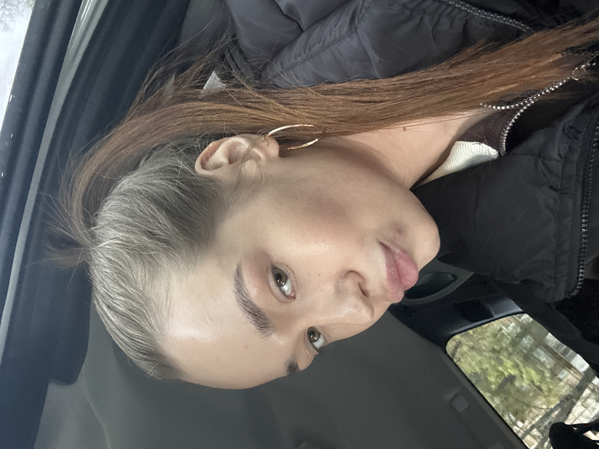 A woman with light skin, light brown and graying hair pulled back, and green eyes is sitting in a car. She is wearing a black puffy jacket and gold hoop earrings. The car's interior is visible, as is a glimpse of trees outside the window, likely in Chaikovsky, Russia. The photo is taken at an angle.