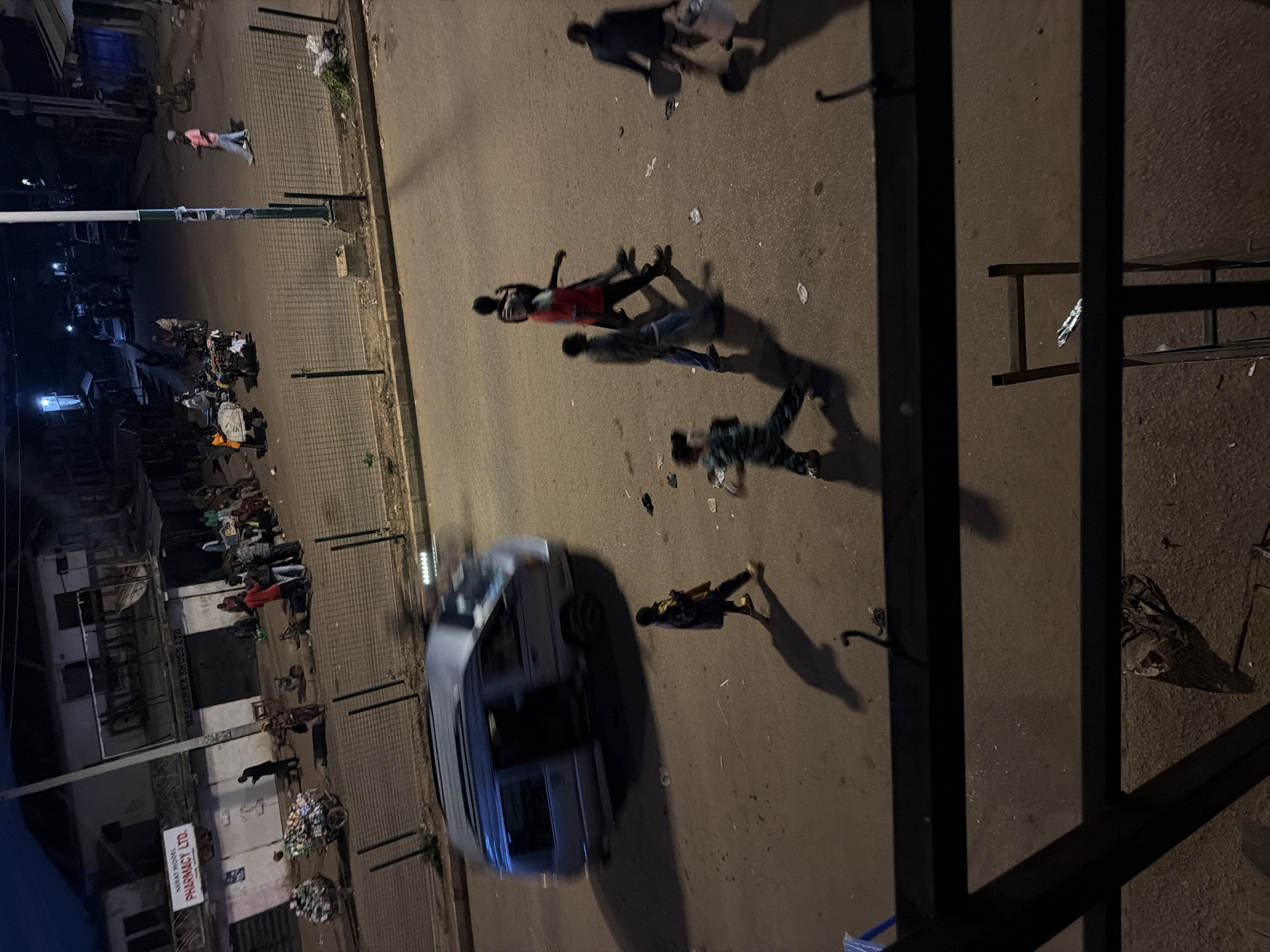The image depicts a nighttime street scene in Bukuru, Nigeria. A silver minivan is captured in motion, appearing blurred due to a long exposure. Several individuals are visible walking on the street and sidewalk. On the left side of the street, a building with a sign reading "NEBAT MODEL PHARMACY LTD." is present. Two carts loaded with what appear to be bottled goods are also on the left. On the right side of the street, a few people are seen near a building, and some trash or debris is scattered on the ground. A wire fence runs along the edge of the street. The overall lighting suggests it is nighttime or dusk.