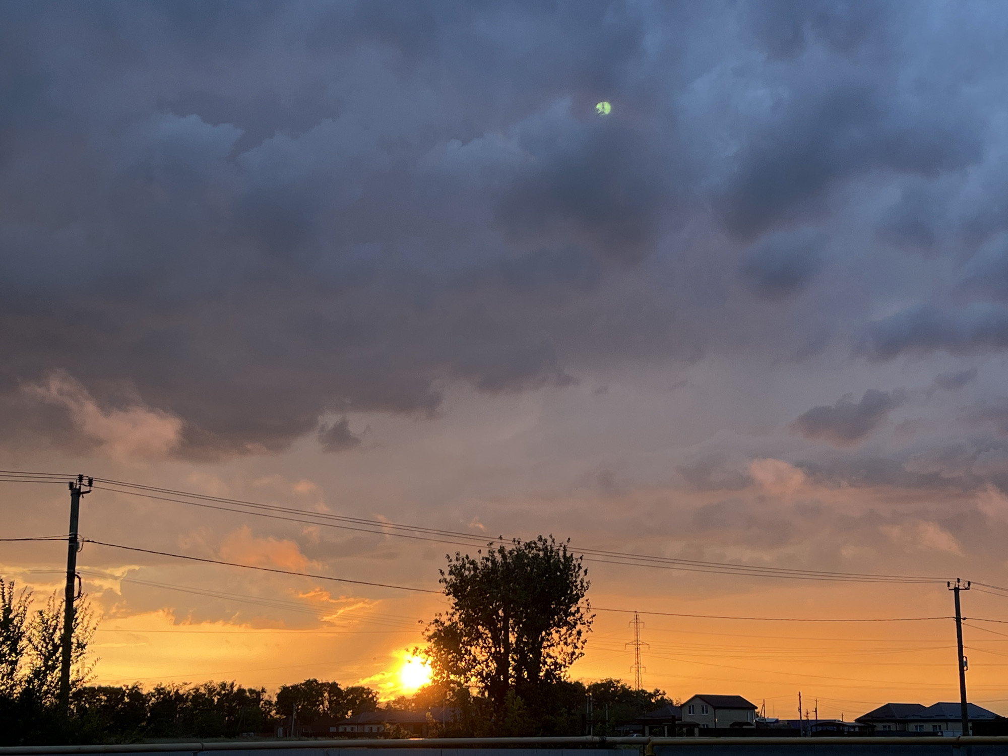 This is an outdoor, daytime, eye-level, full-shot photograph taken in the Taganrog City District, Russia. The image captures a dramatic sunset scene. The sky is filled with a gradient of colors, transitioning from a deep, stormy gray at the top to vibrant oranges and pinks near the horizon. Dark, heavy clouds dominate the upper portion of the sky, while lighter, more wispy clouds are illuminated by the setting sun.
In the middle ground, a silhouette of trees forms a dense line across the horizon, with a prominent, leafy tree standing out in the center. The sun is partially obscured by this tree and is emitting a bright, intense light that creates a halo effect.
Several utility poles with wires are visible throughout the frame, indicating the presence of human infrastructure. In the background, the silhouettes of houses and a tall power pylon are discernible against the bright sky. A faint, round, yellowish-green object, possibly a balloon or a frisbee, is visible in the upper right quadrant of the sky. The foreground shows a dark, horizontal railing or barrier.