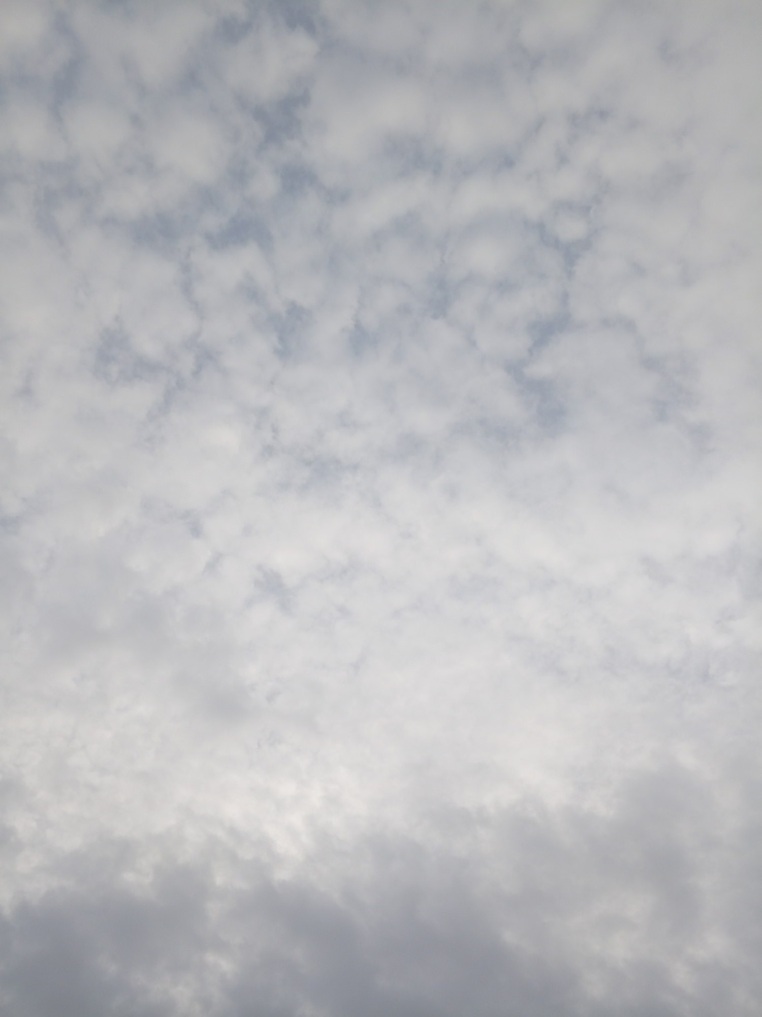 The image shows a cloudy sky. The clouds in the upper portion of the image are smaller and more scattered, revealing glimpses of blue sky. The clouds in the lower portion of the image are denser and darker, forming a thick layer. There are no people, subjects, objects, actions, or interactions visible in the image. The location of the image is Mbribit, Nigeria.