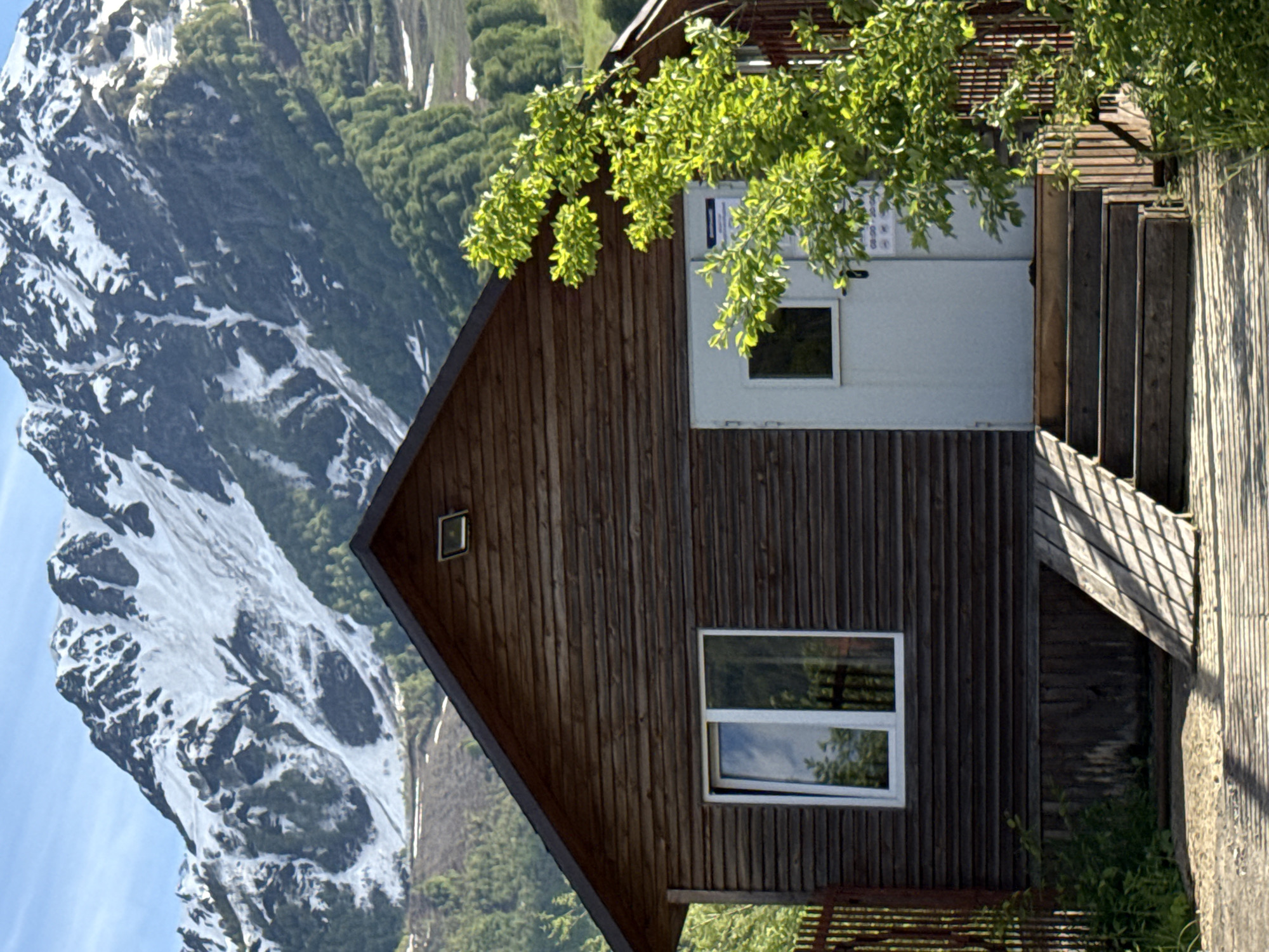 A wooden house or cabin with a steep gabled roof stands prominently in the foreground, set against a dramatic backdrop of snow-capped mountains. The house is constructed from vertical dark brown wooden planks. On its main face, a rectangular white-framed window reflects the sky and surrounding trees. A smaller square window is visible higher up near the roof's peak. To the right, a light-colored, possibly white or light grey, door with a small window is accessible via wooden steps that lead up from the right side.
Lush green foliage from a tree hangs over the upper right side of the house, casting dappled light and shadows. On the wall next to the door, several light-colored signs or posters are affixed. While most text is unreadable, some faint characters and numbers, including what appears to be "10:00 - 20:00" or similar time notations, can be vaguely discerned on these signs.
The background is dominated by rugged mountains, heavily dusted with snow, interspersed with dark rock faces and patches of green coniferous trees. The sky above is clear and bright blue. The scene is bathed in strong daylight, indicating it is daytime with sunny weather. No people or specific activities are taking place. The image depicts a tranquil, scenic mountain setting, consistent with the location of Estosadok, Russia.